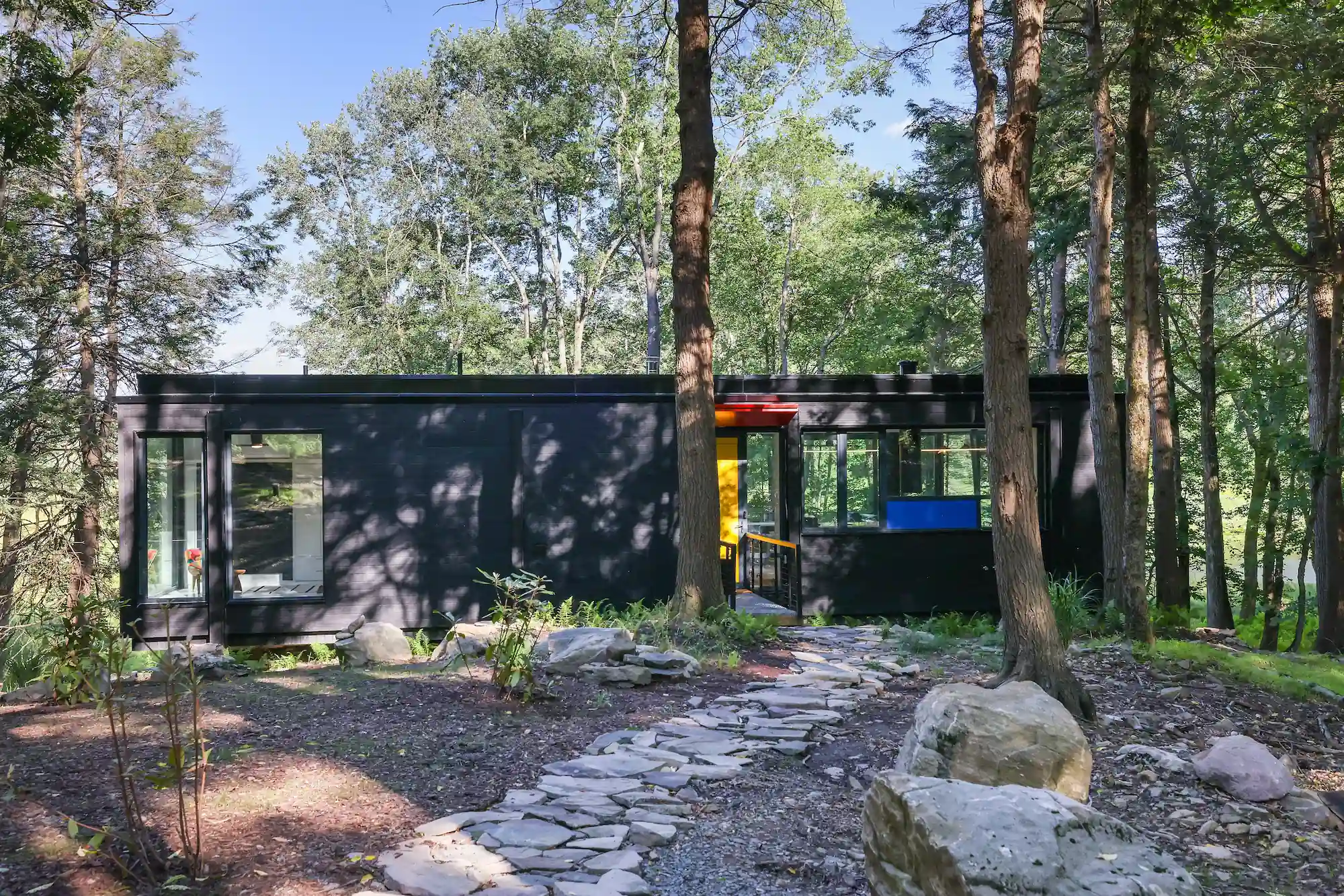 Stone path leading to the charred wood exterior, its colorful door accents visible through the forest.