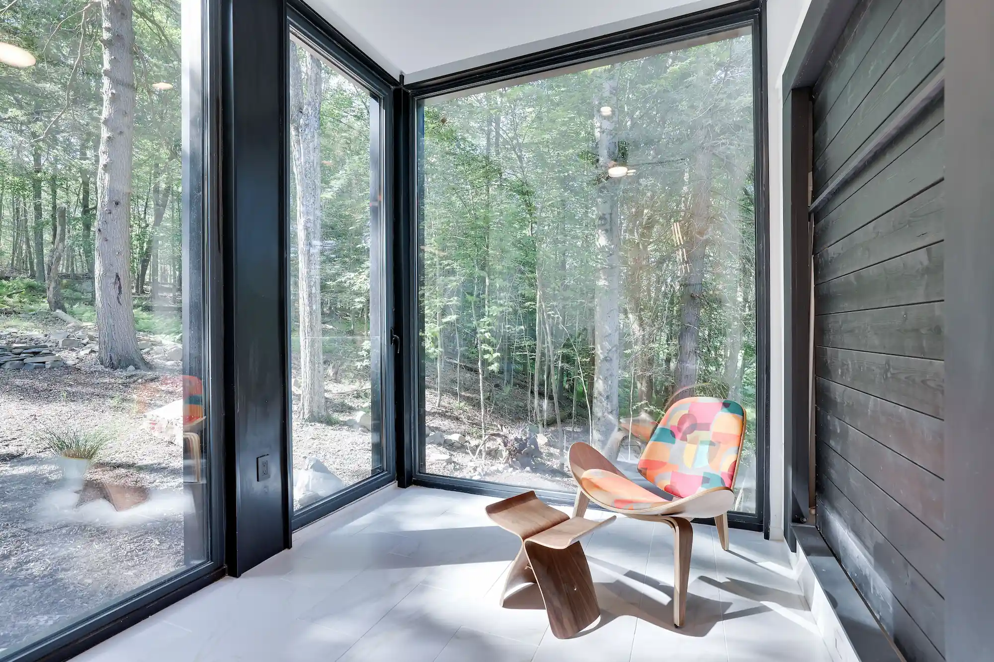 Corner glass nook with a mid-century lounge chair framed by triple-glazed windows and wooded views.
