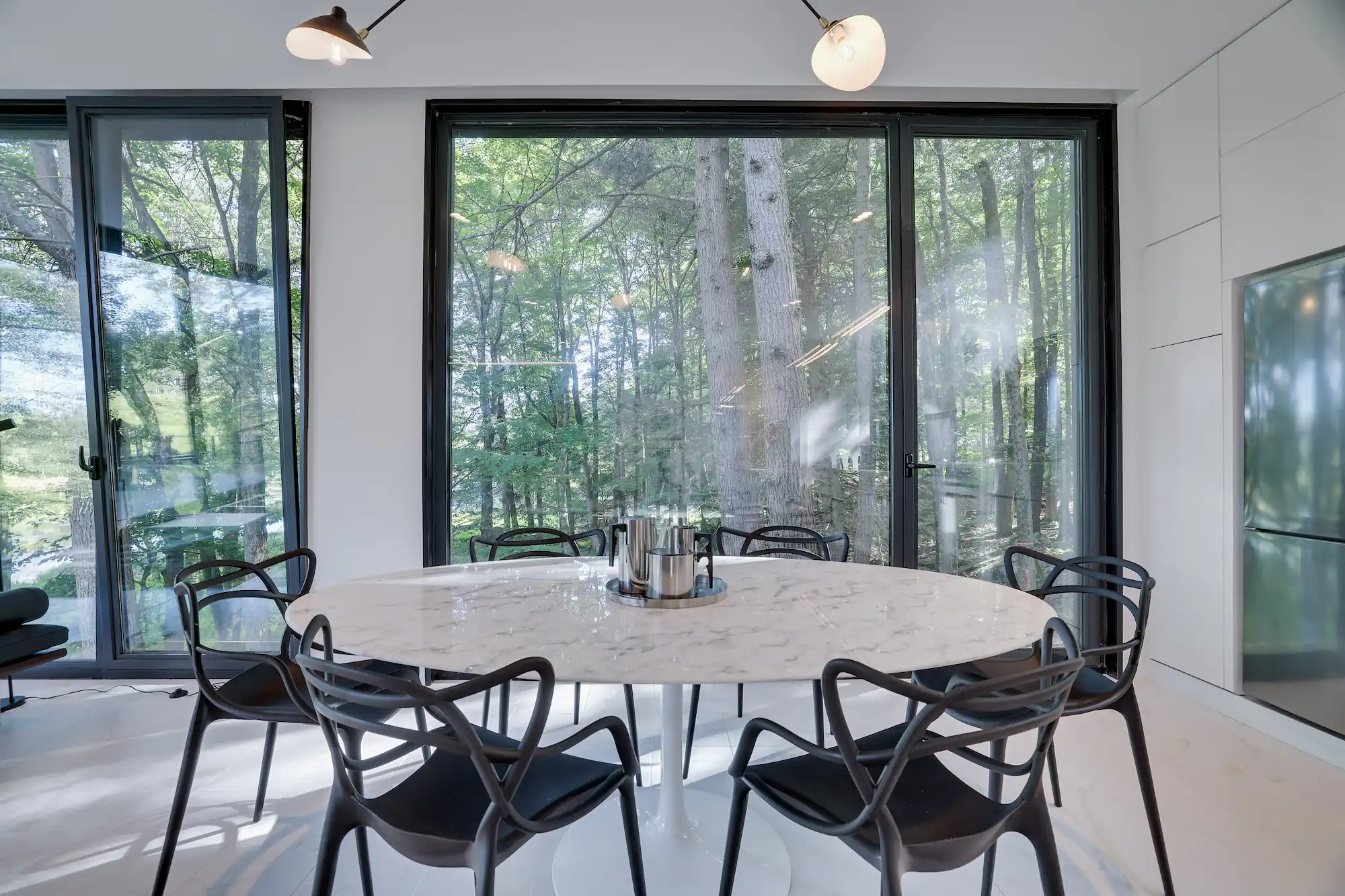 Dining table and mid-century chairs surrounded by floor-to-ceiling glass overlooking the forest.