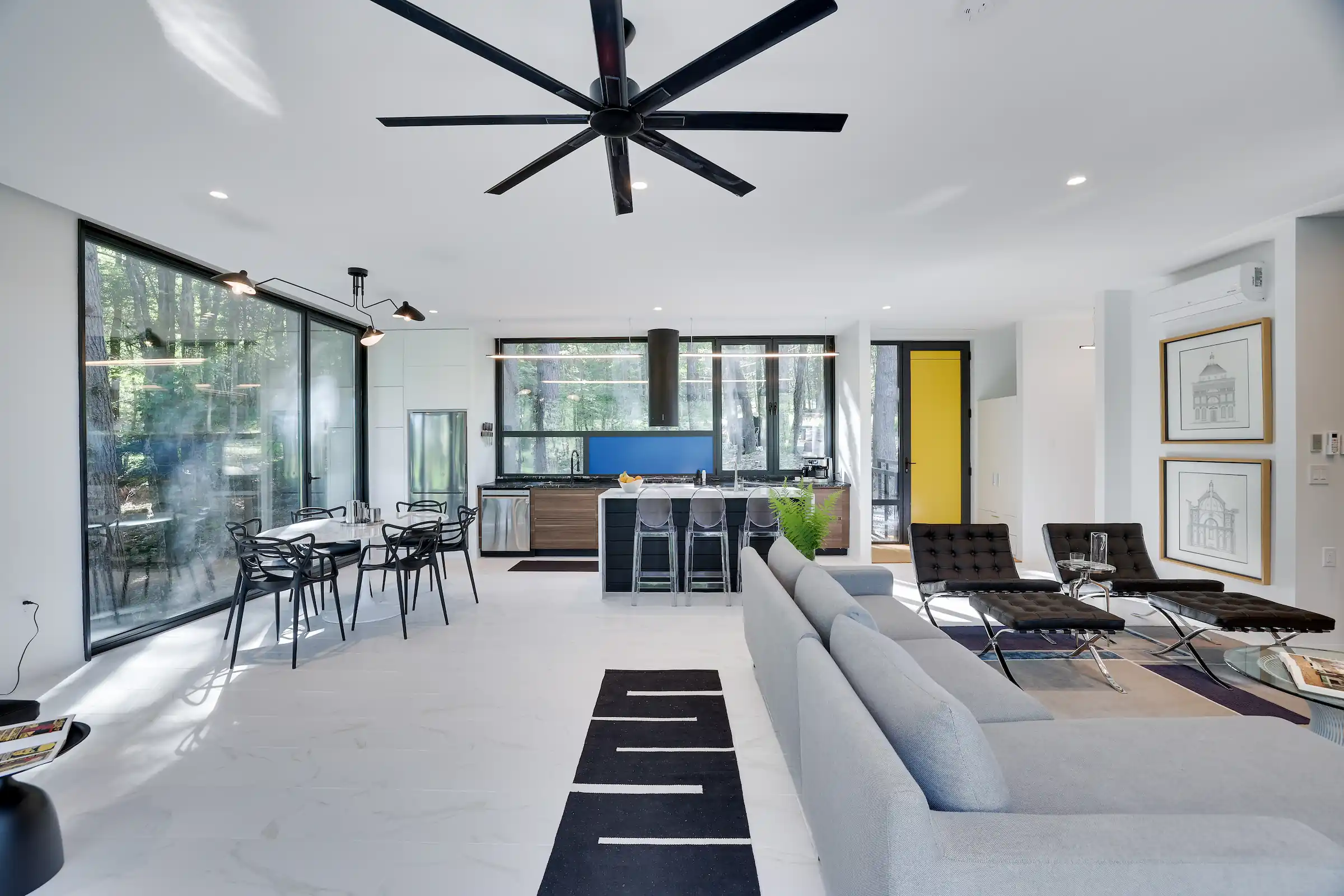Open-plan living, dining, and kitchen area wrapped in floor-to-ceiling glass with forest views on both sides.