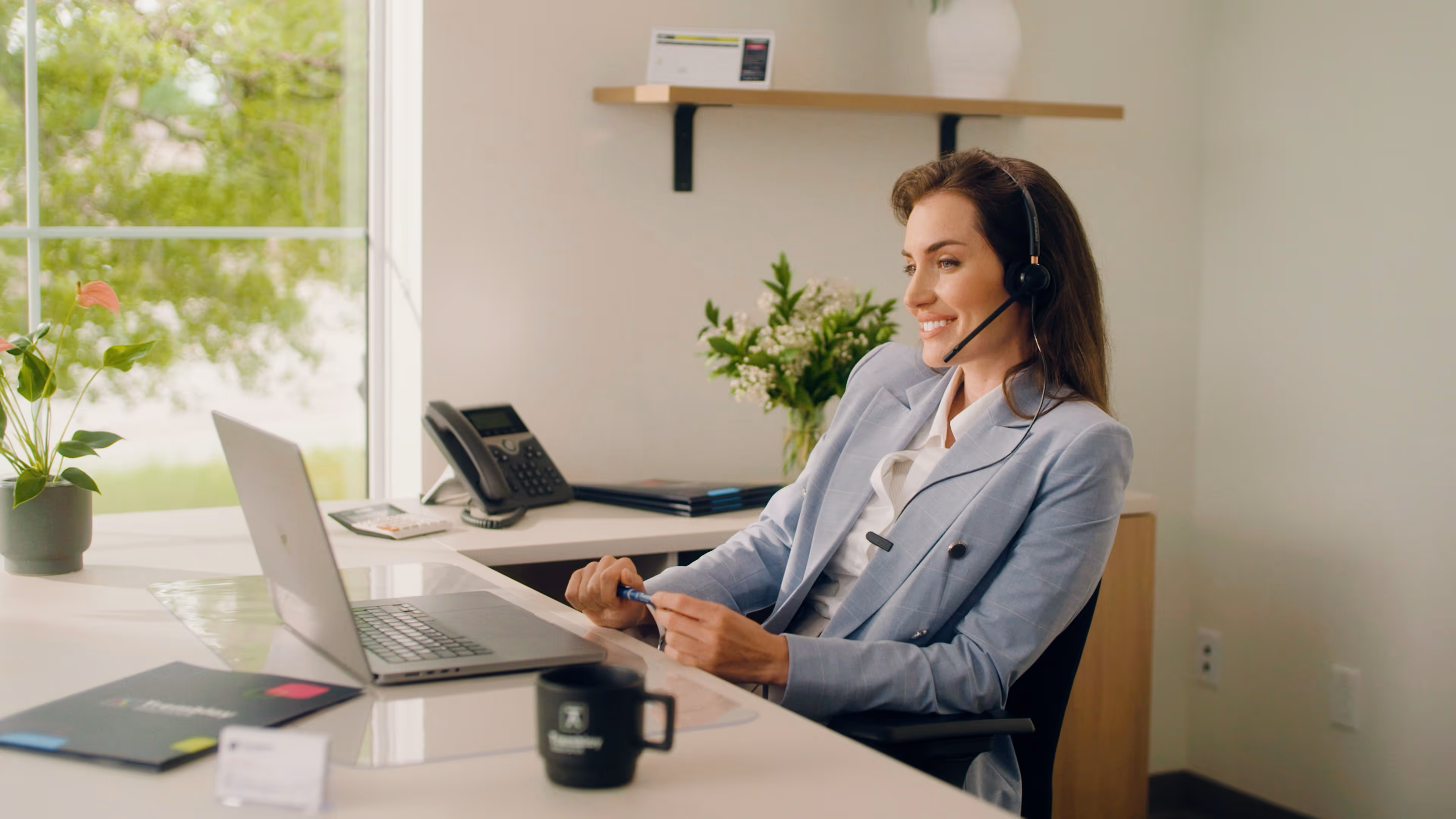 Femme souriante en veste bleue portant un casque, travaillant sur un ordinateur portable dans un bureau lumineux avec une plante et un téléphone de bureau.
