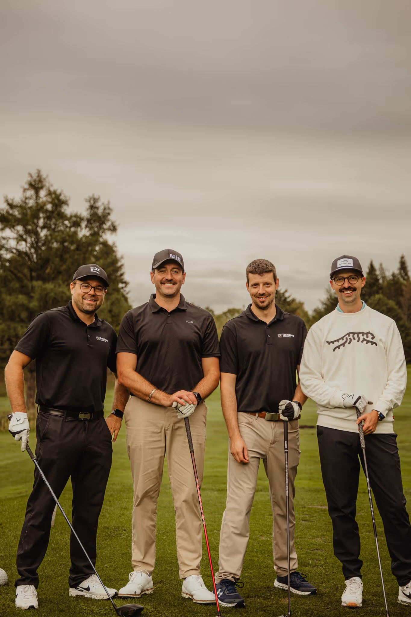 Quatre hommes souriants posant sur un terrain de golf avec des bâtons de golf, habillés en tenue décontractée de golf.