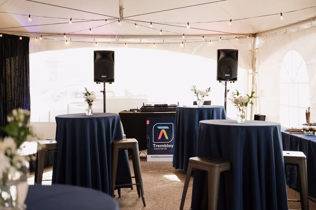 Salle d'événement avec des tables hautes recouvertes de nappes bleues et des tabourets métalliques, un stand DJ avec deux haut-parleurs et des bouquets de fleurs.