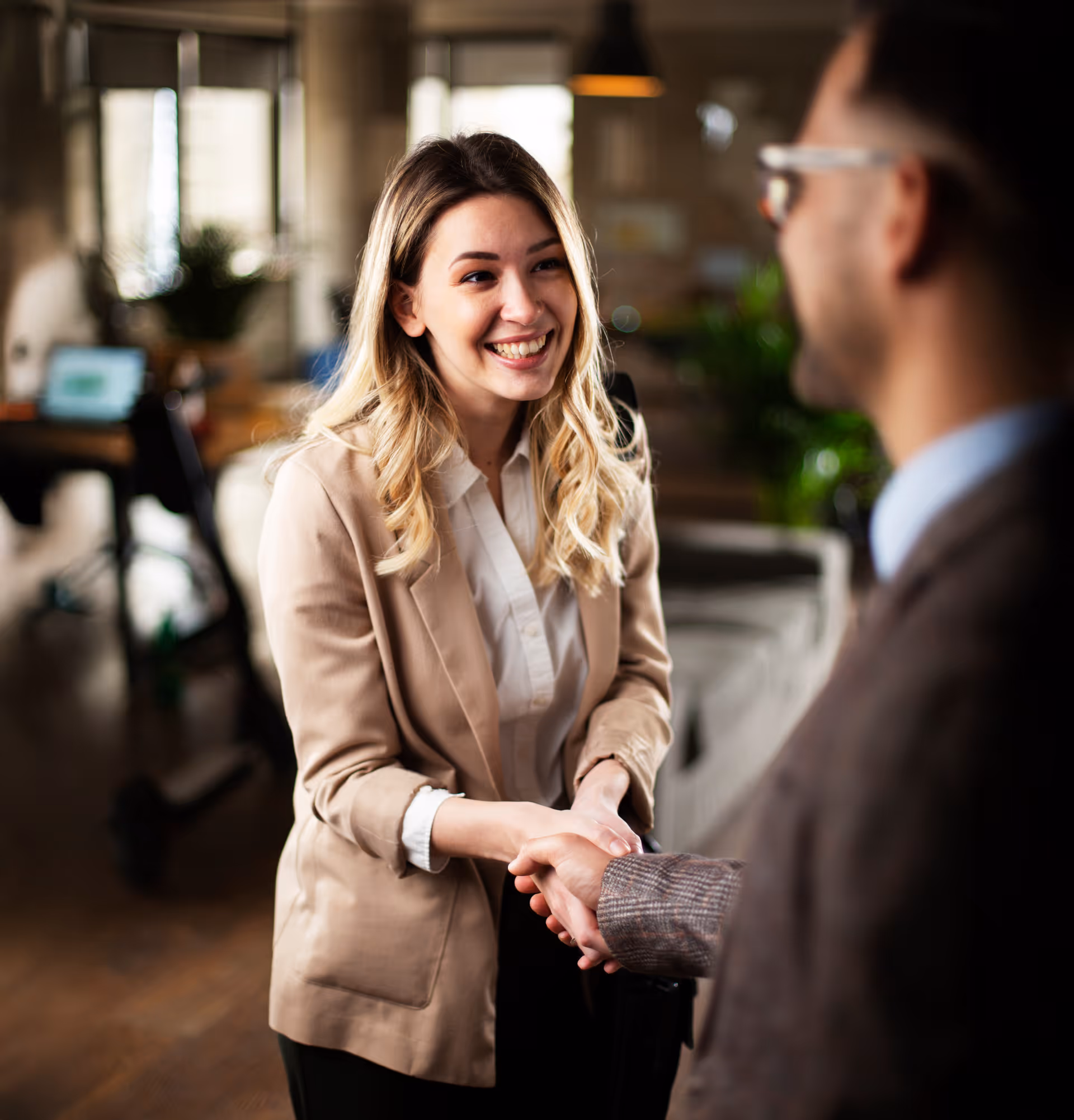 Une femme souriante en veste beige serre la main d'un homme lors d'une rencontre professionnelle dans un bureau.