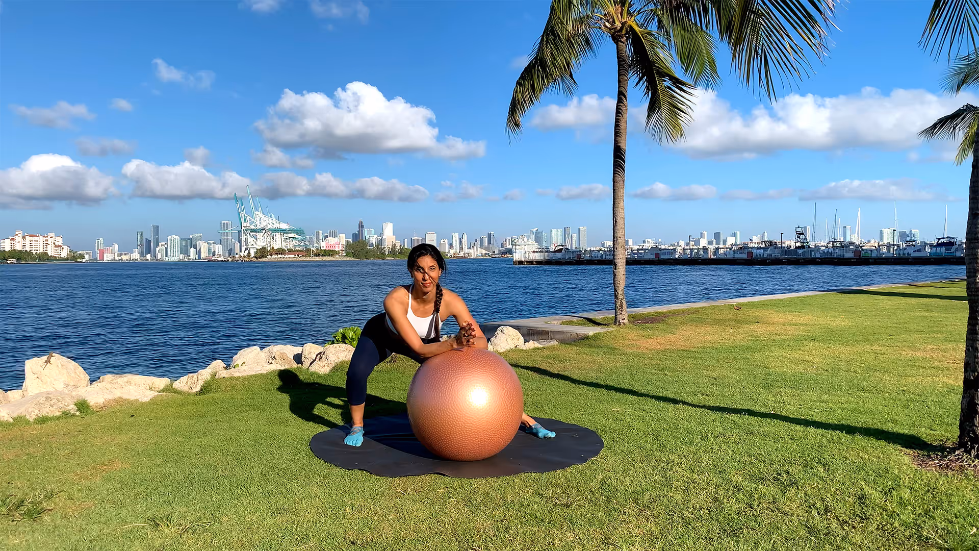 Woman doing a stability ball Pilates exercise outdoors by the ocean with a city skyline in the background.