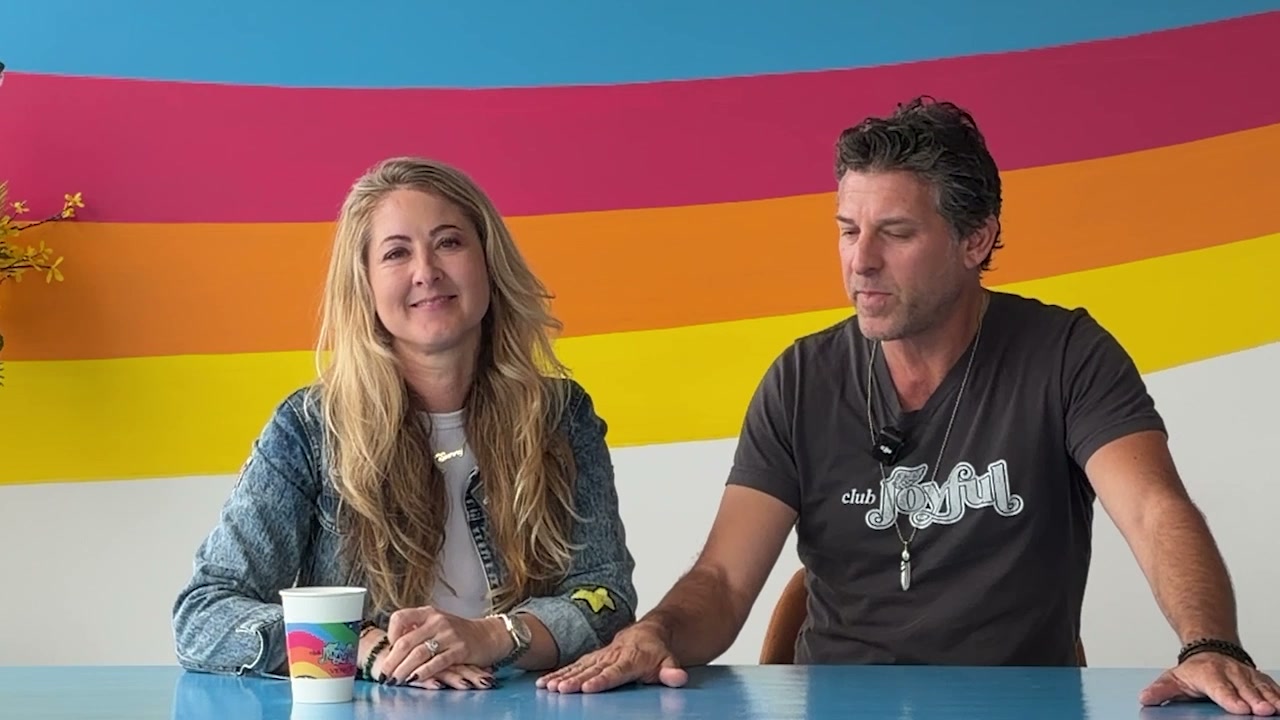 Erika and Scott of Club Joyful seated in front of the rainbow mural in Venice Beach, a rainbow latte on the table