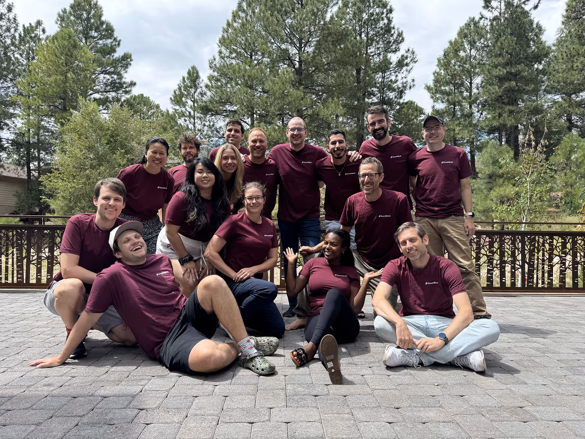 Group photo of sixteen diverse people wearing matching maroon shirts outdoors on a stone patio with trees in the background.