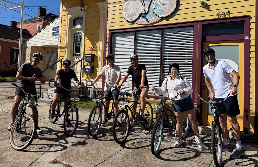 Group of six cyclists wearing helmets standing with their bikes in front of a yellow building with a bicycle-themed sign.