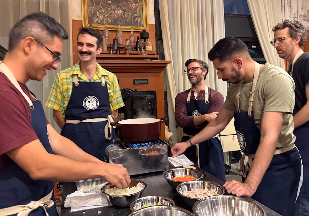 Five men wearing aprons preparing ingredients and cooking together in a kitchen with a pot on a stove.