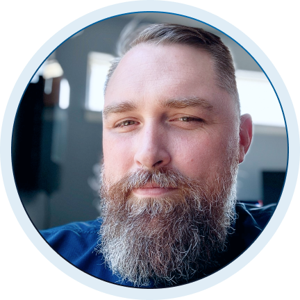 Close-up portrait of a man with a full beard and mustache, light eyes, and combed-back hair, in a softly lit indoor setting.