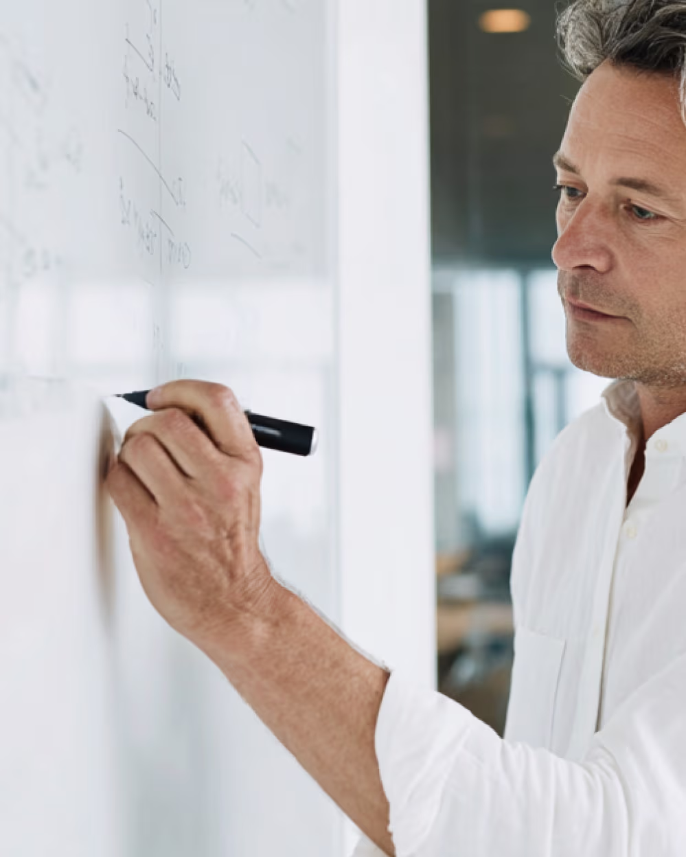 Man writing with black marker on a whiteboard filled with faint notes in an office setting.