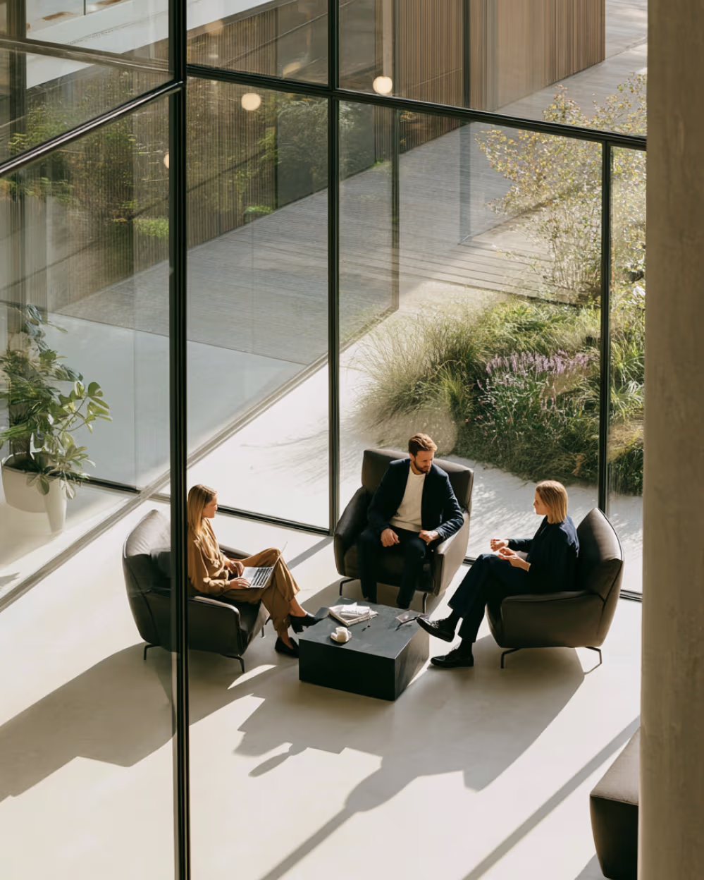 Three people seated in a modern glass-walled room having a discussion around a black coffee table.