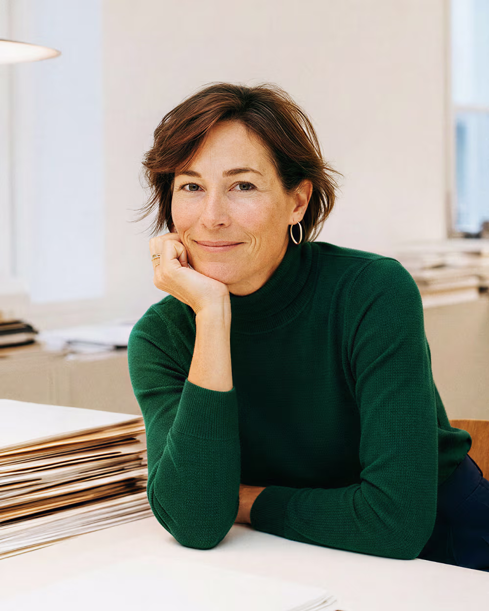 Woman in green sweater leaning on a table with documents, smiling gently at camera.