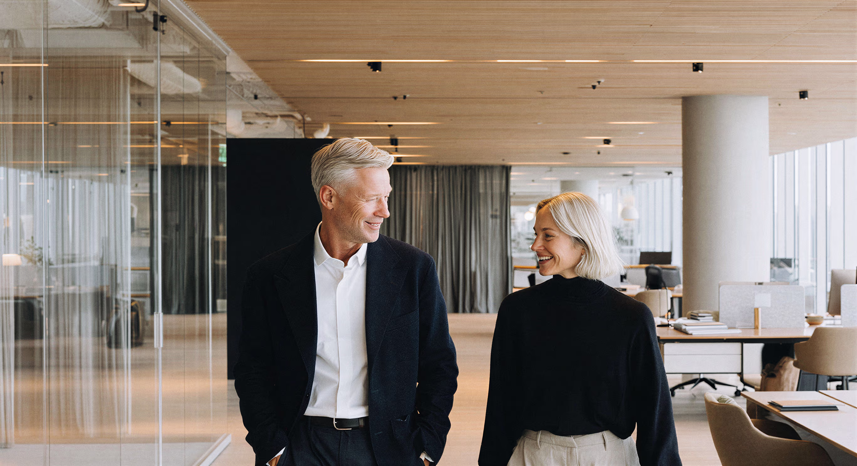 Man and woman in business attire smiling and walking in a modern office space with desks and glass walls.