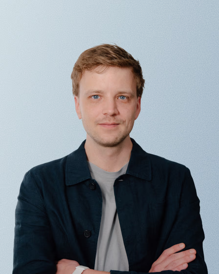 Young man with light brown hair and blue eyes wearing a black jacket, arms crossed, smiling slightly.