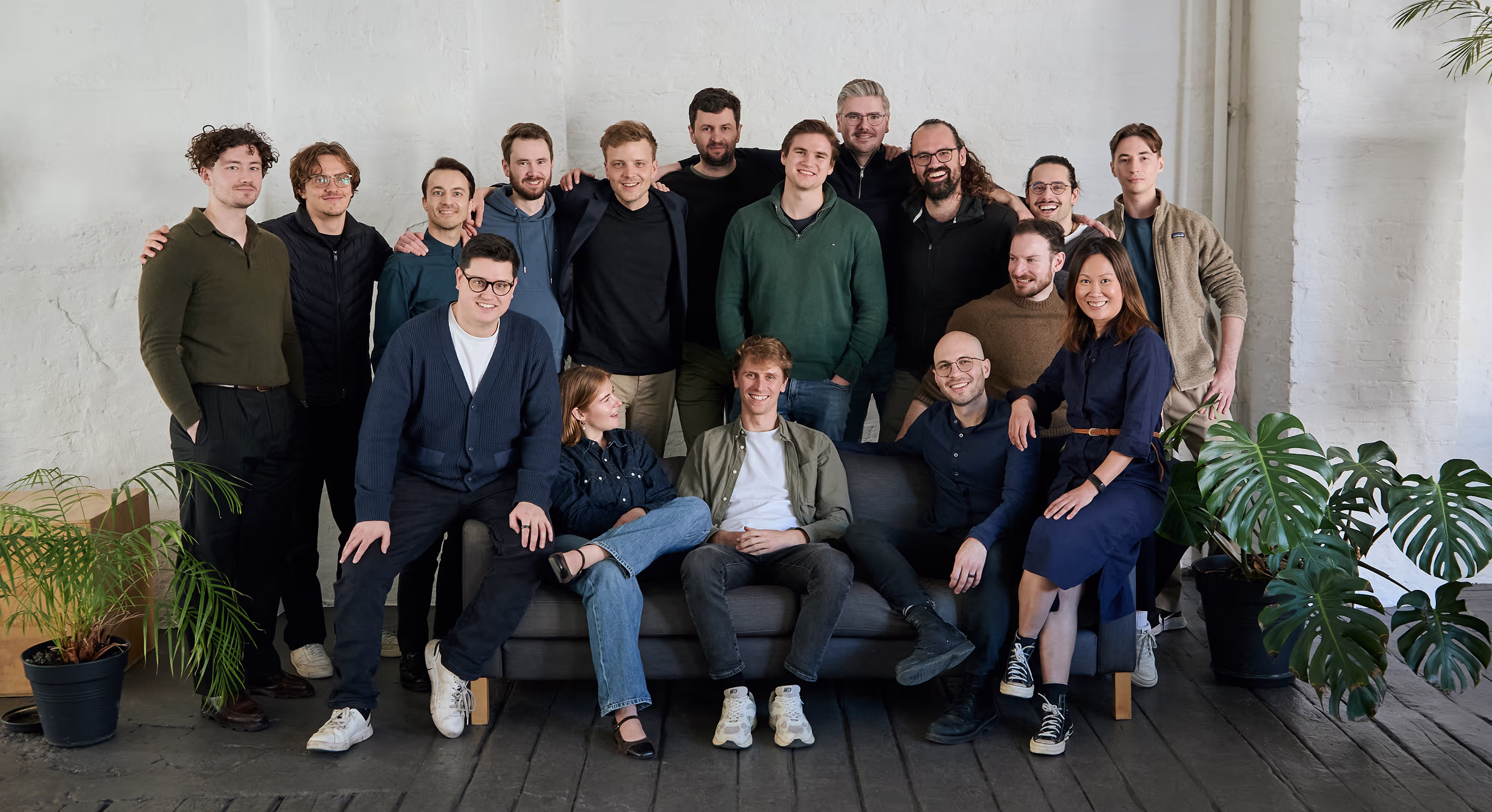 Group of 17 diverse coworkers smiling and posing casually in a modern office with plants and a gray sofa.