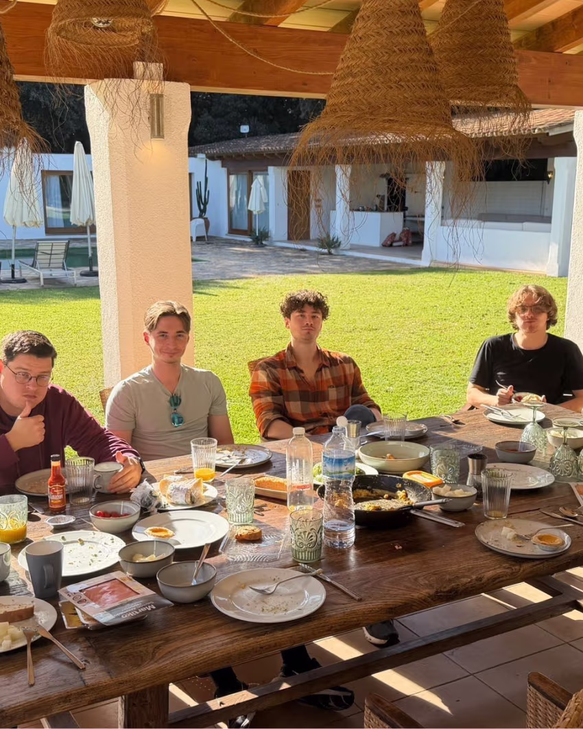 Four men sitting outdoors at a wooden table with plates and glasses after a meal in a sunny garden.