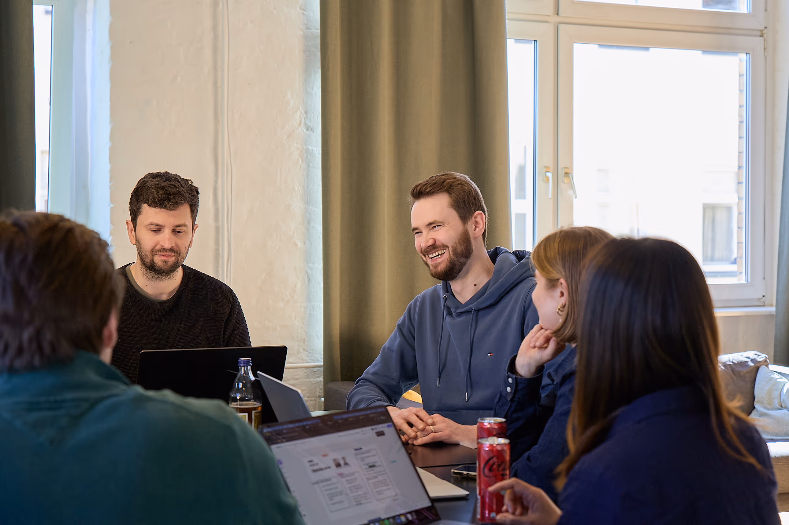 Five people in casual clothes sitting around a table with laptops, chatting in a bright room.