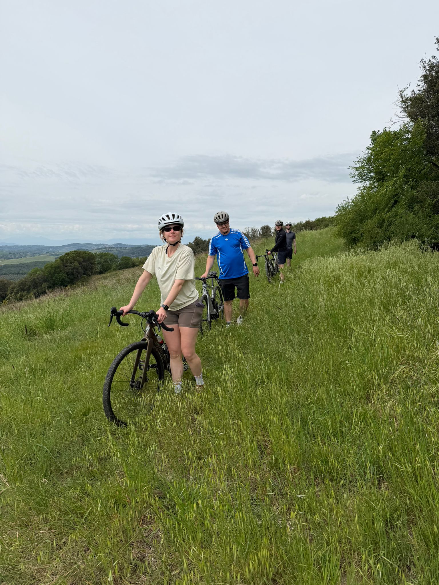 Four cyclists walking with bikes through tall grass on a countryside trail under a cloudy sky.