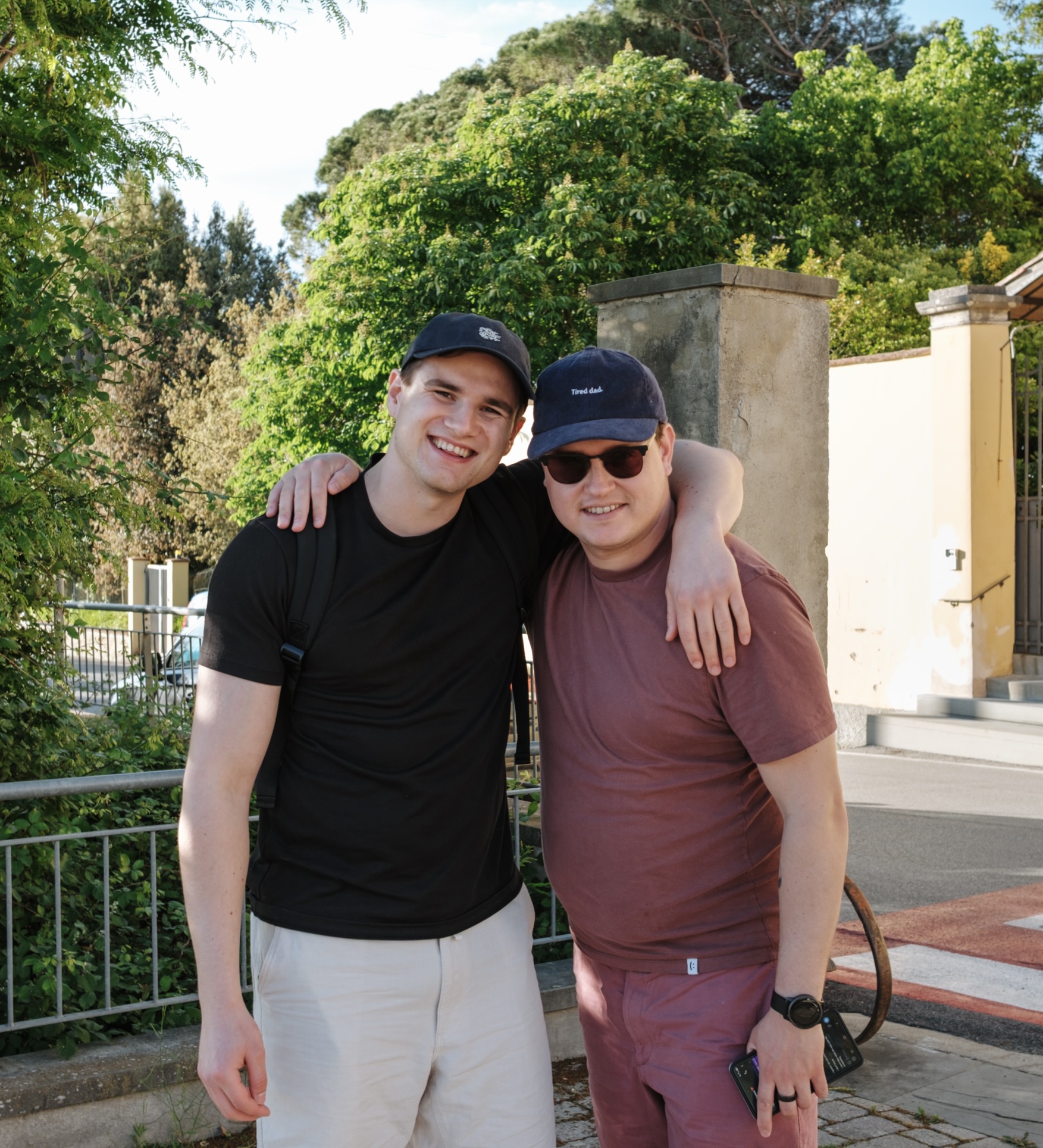 Two men outdoors smiling, one in black shirt with arm around man in maroon shirt and cap.