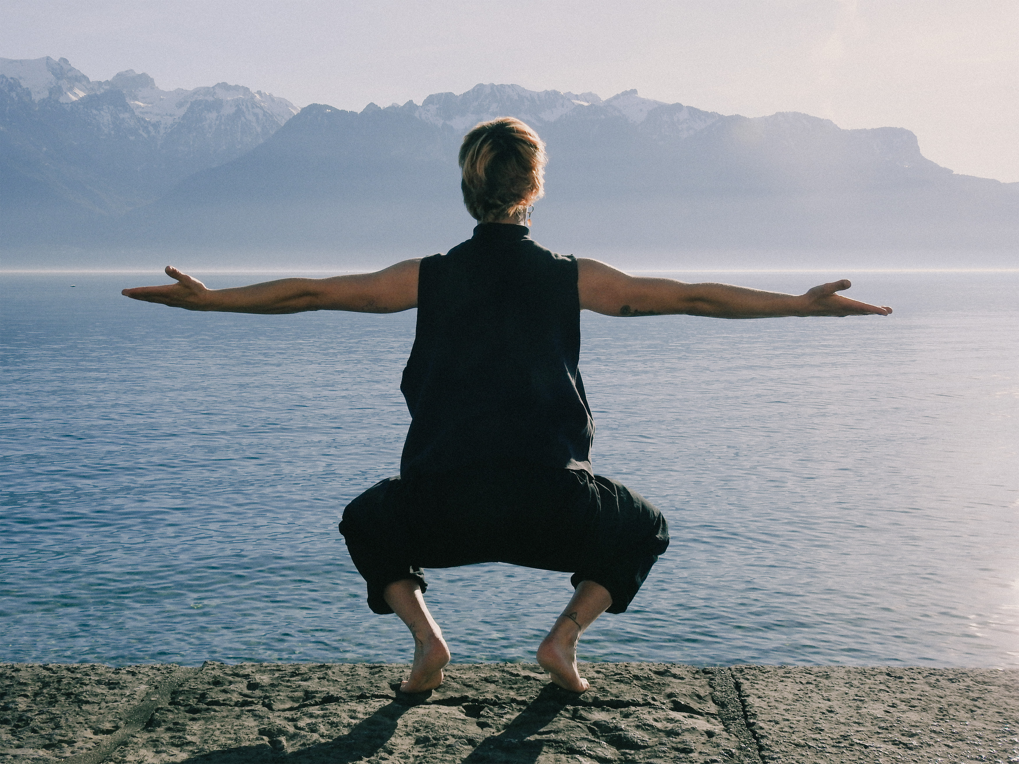 Person squatting barefoot on a stone surface near a large body of water with arms extended, mountains in the background.