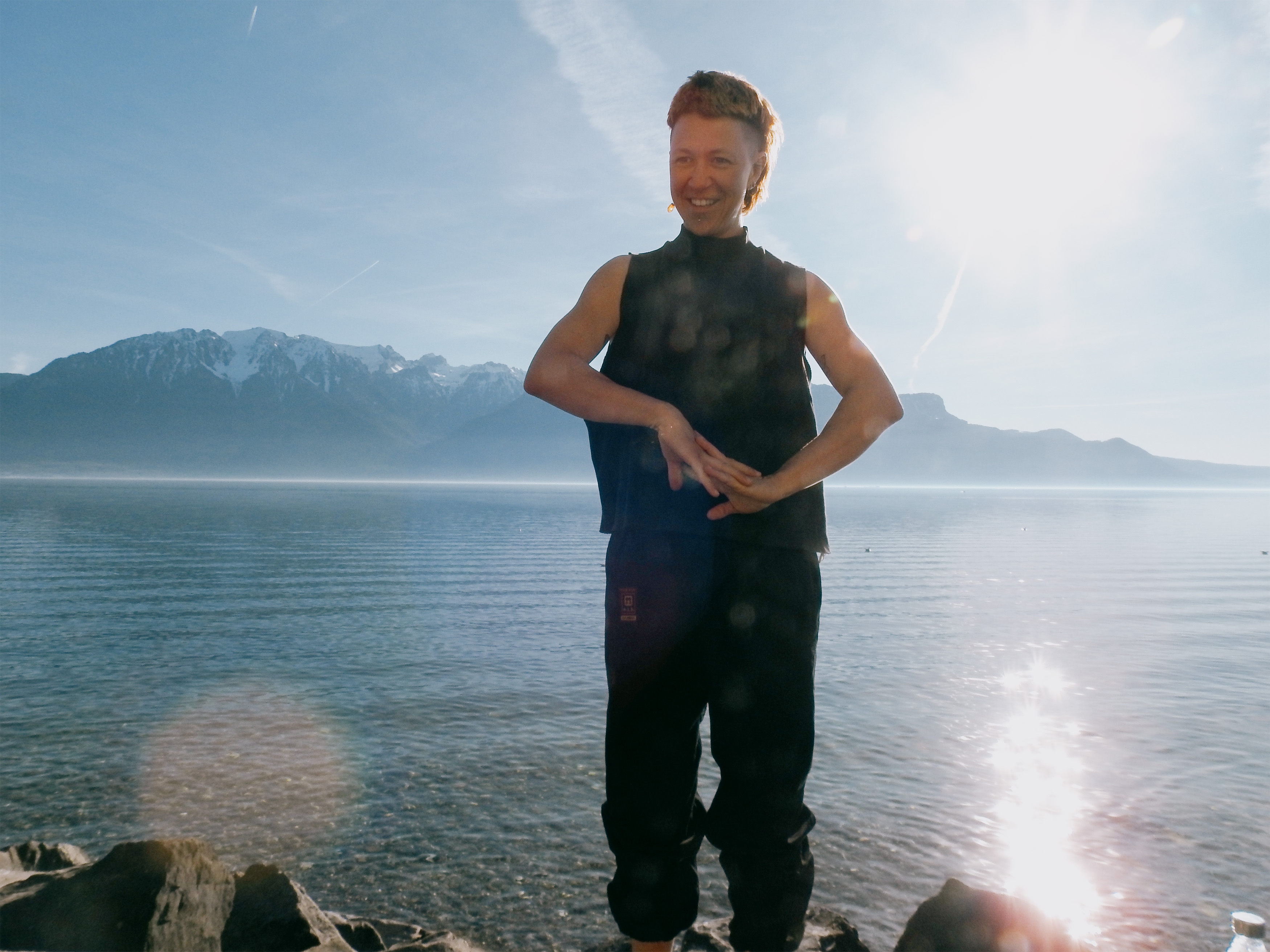 Person standing on rocky shore with a calm lake and snow-capped mountains in the background under a clear blue sky.