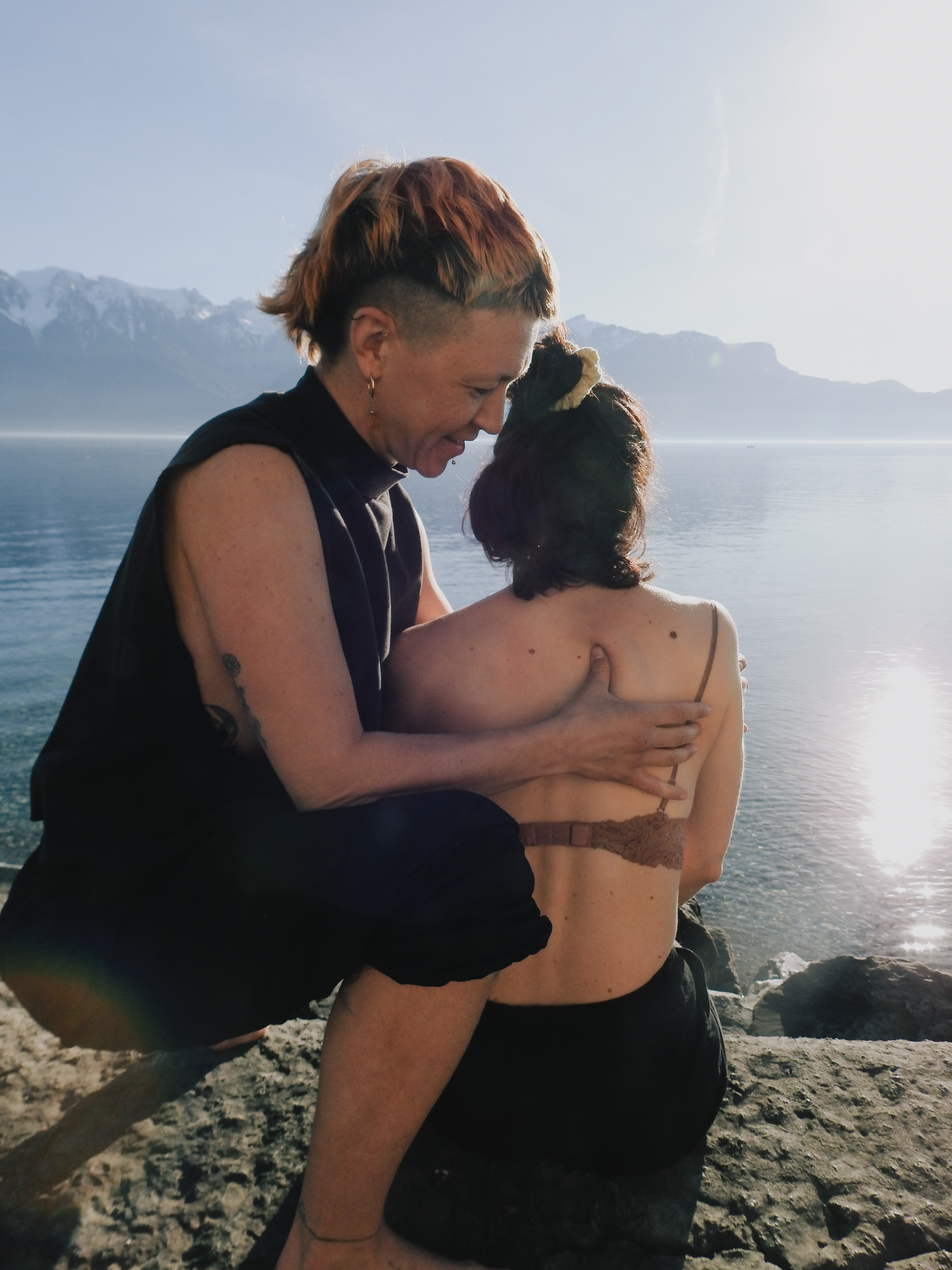 Two people sitting by a lake with mountains in the background, one person embracing the other.