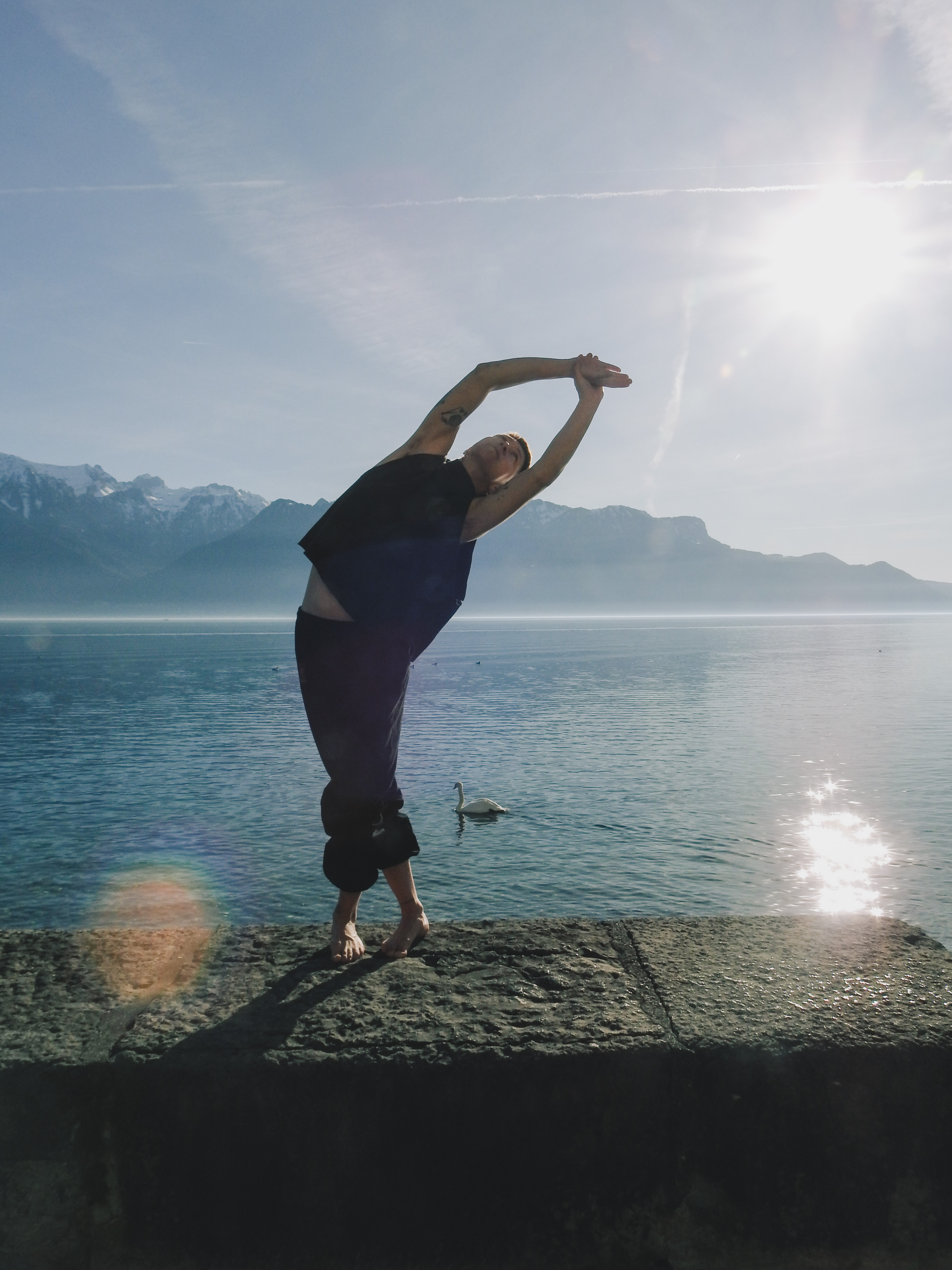 Person stretching barefoot on a stone ledge by a calm lake with mountains and a swan in the background under a sunny sky.