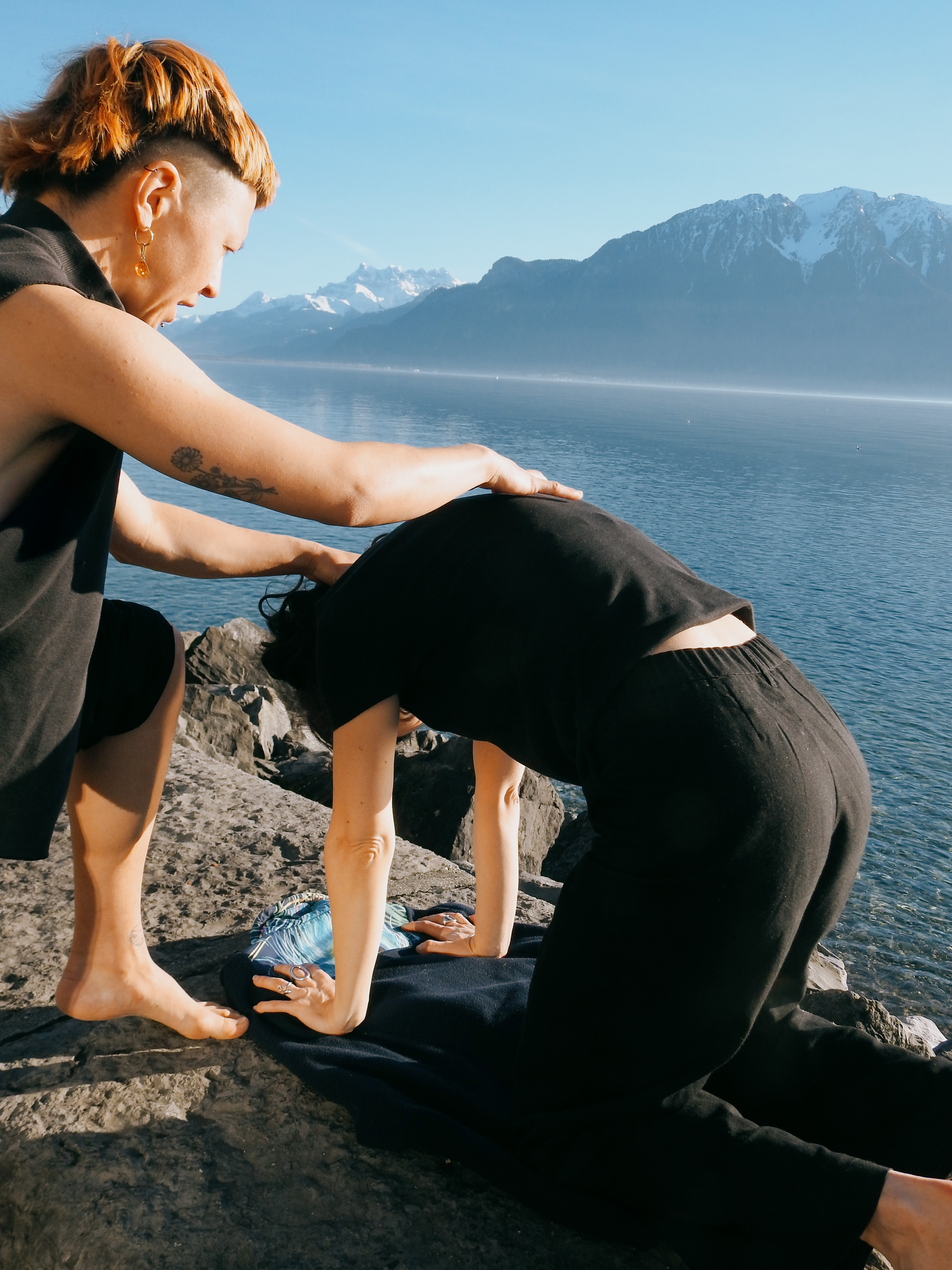 Person performing assisted stretching on another person kneeling on a rocky shore by a lake with mountains in the background.