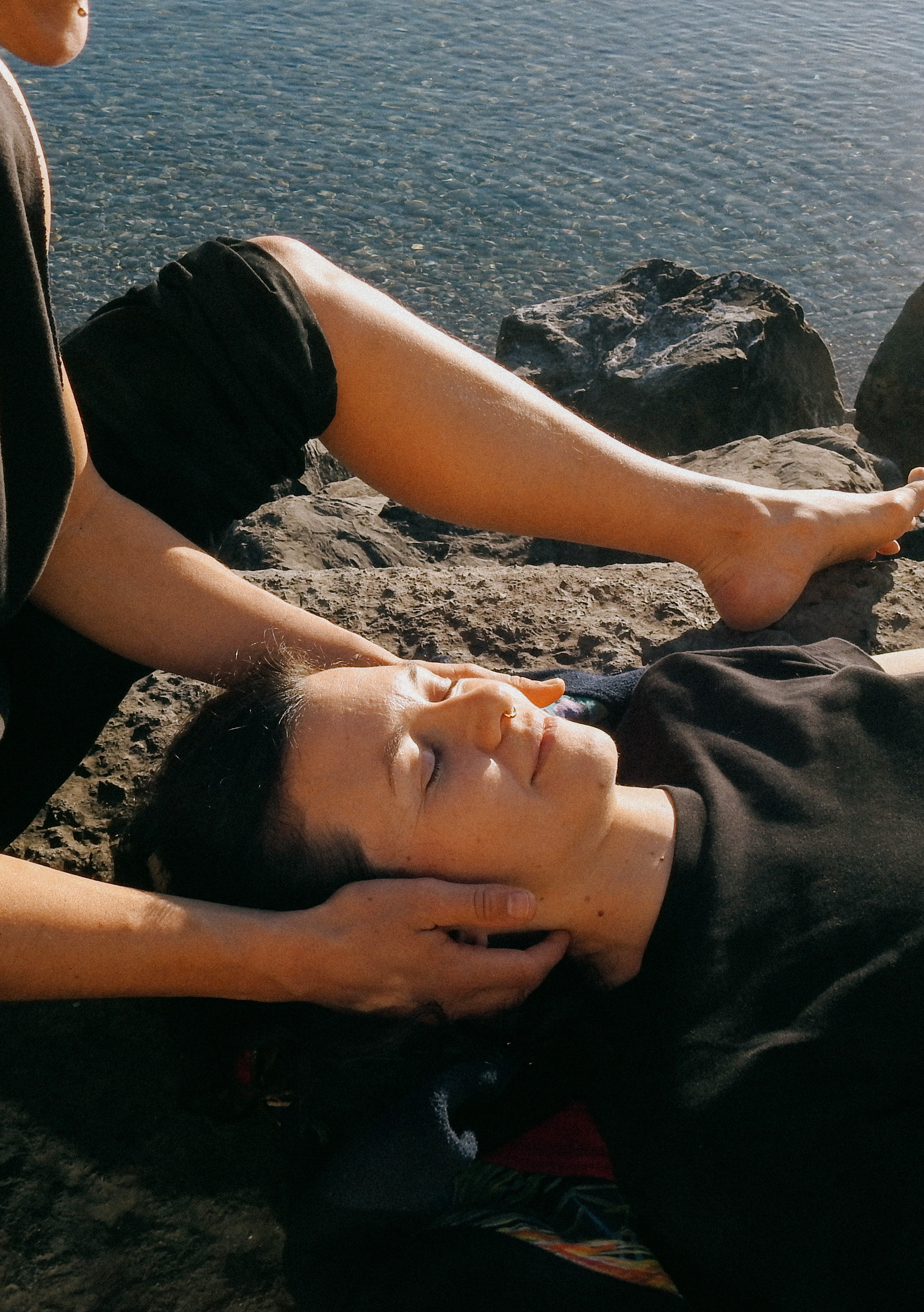 Person receiving a gentle head massage while lying near calm water and rocks.