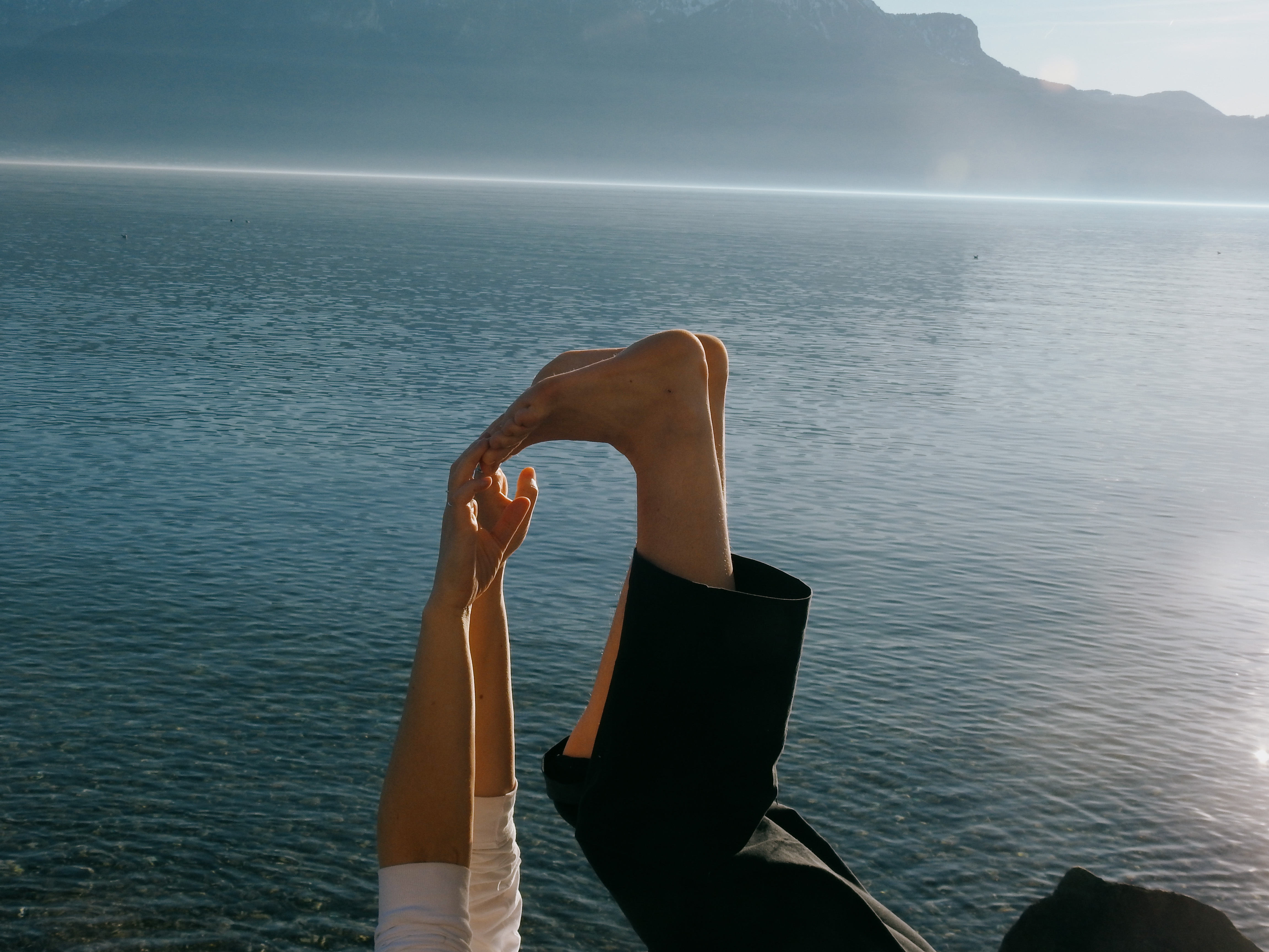 Person by a calm lake bending one leg and holding their foot with both hands against a backdrop of misty mountains.
