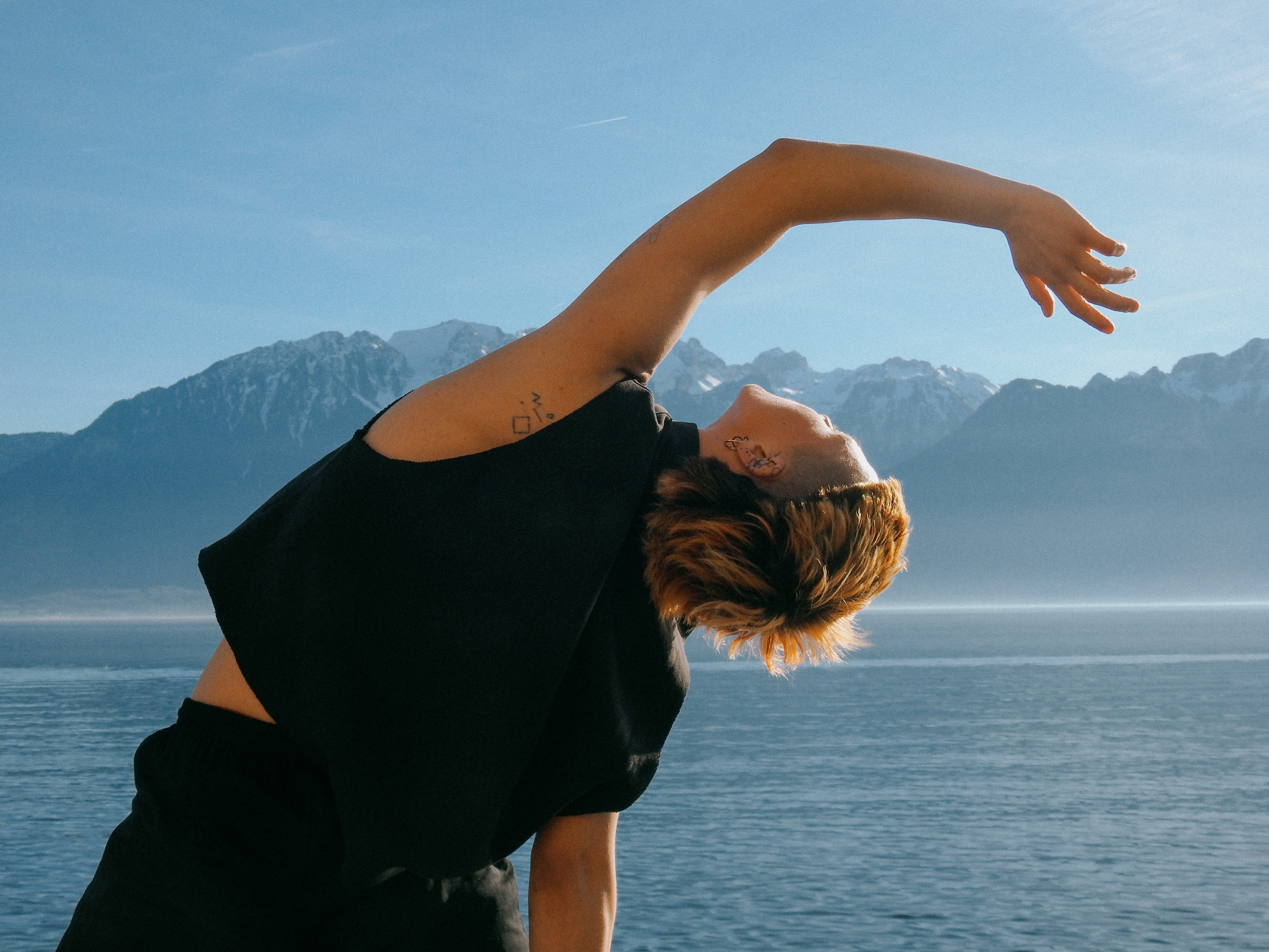 Person with short hair stretching backwards near a calm body of water with mountains in the background.