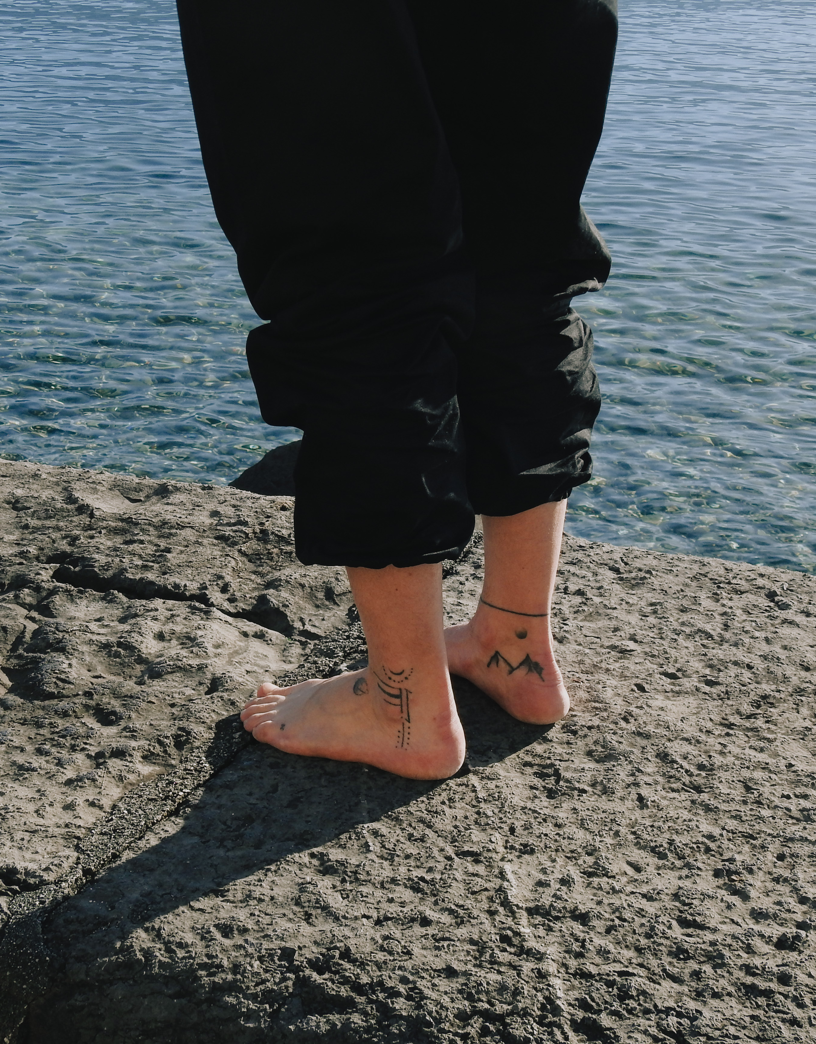 Bare feet with black tattoos standing on rocky shore near clear water.