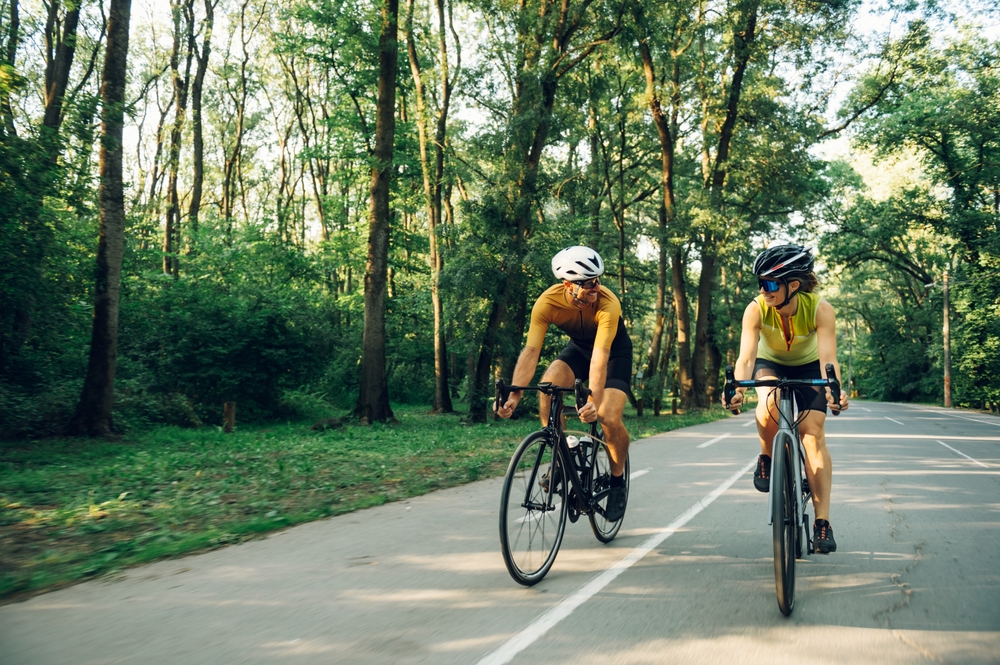 A man and a woman riding their bikes through a countryside road, surrounded by trees.