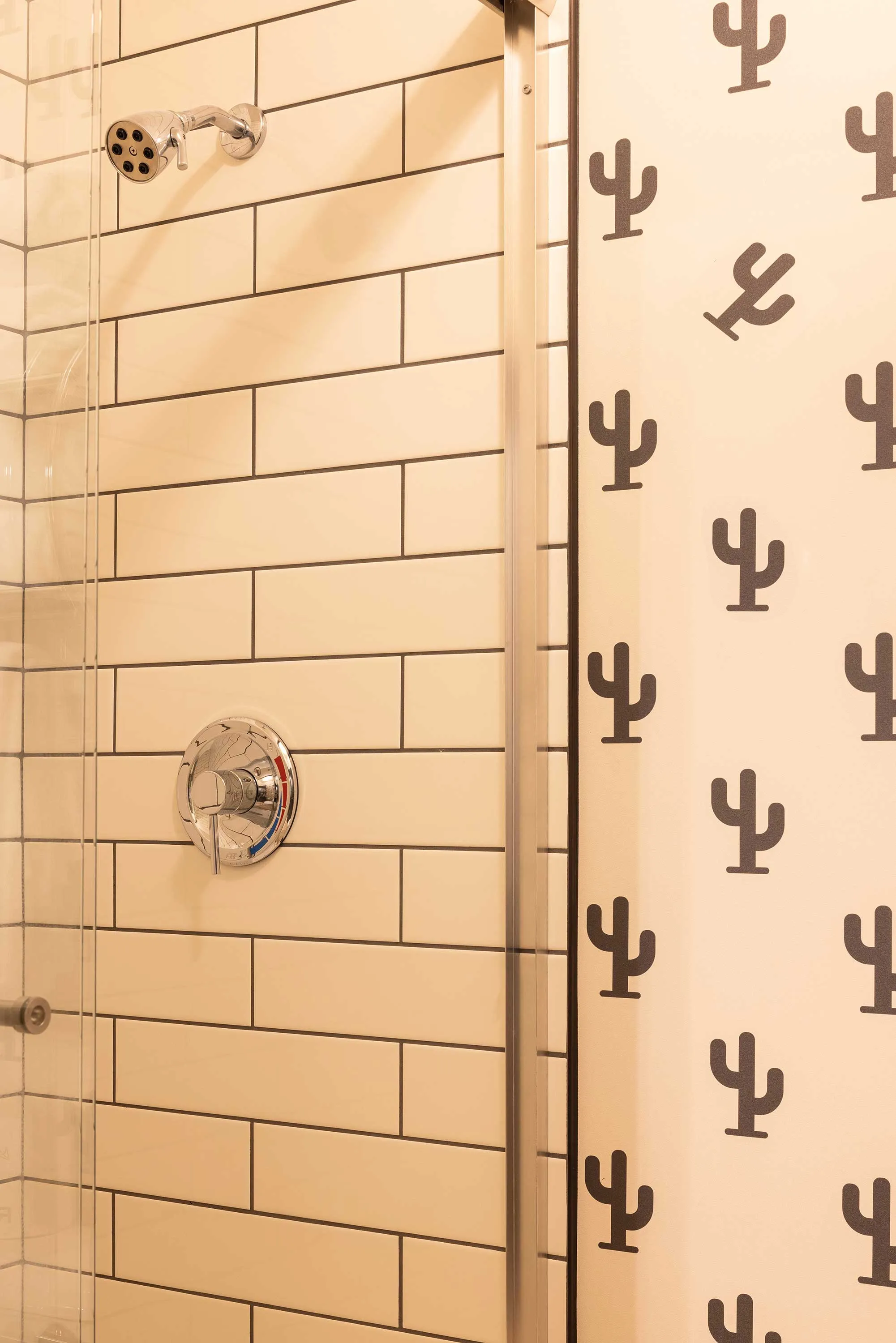 Shower area with white rectangular subway tiles and a chrome showerhead and handle, adjacent to a wall with a cactus pattern. Mountain Modern Motel Sedona, custom surface pattern design