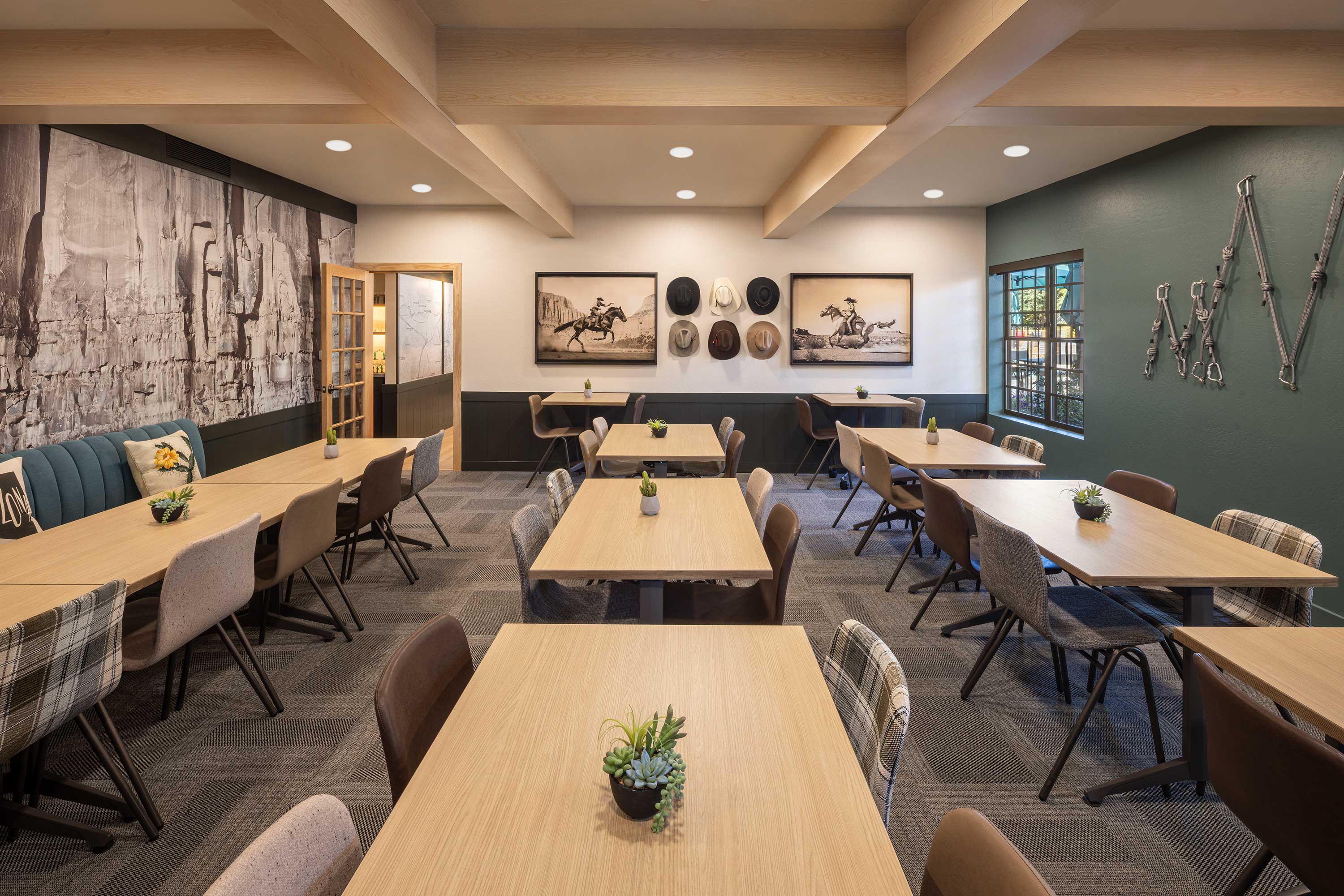 Modern dining room with wooden tables, mixed chairs, cowboy hat wall decor, black-and-white rodeo photographs, and a large rock formation mural. Mountain Modern Motel Sedona