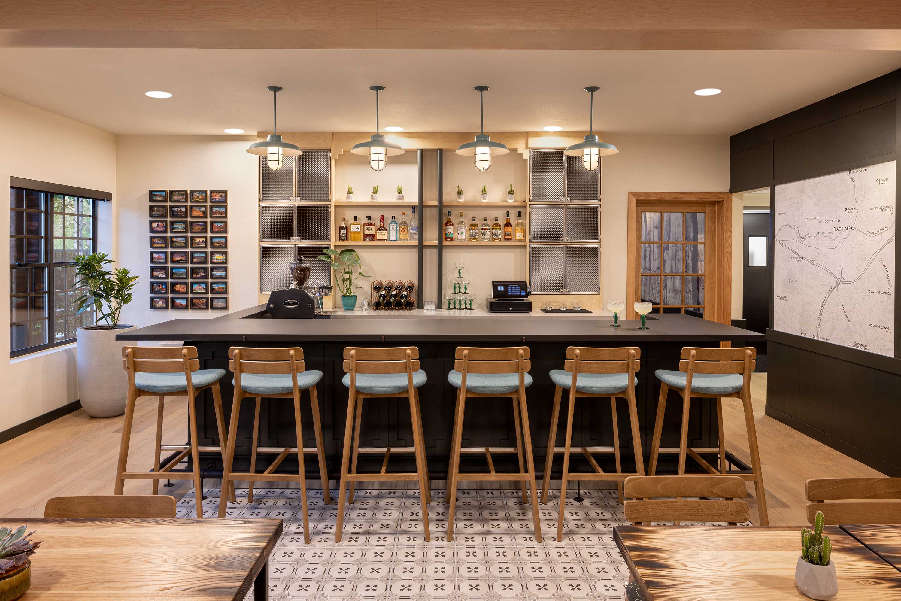 Modern bar interior with six wooden stools, pendant lights, a shelf with liquor bottles, plants, and a large map on the wall. Mountain Modern Motel Sedona, geographic map wall covering