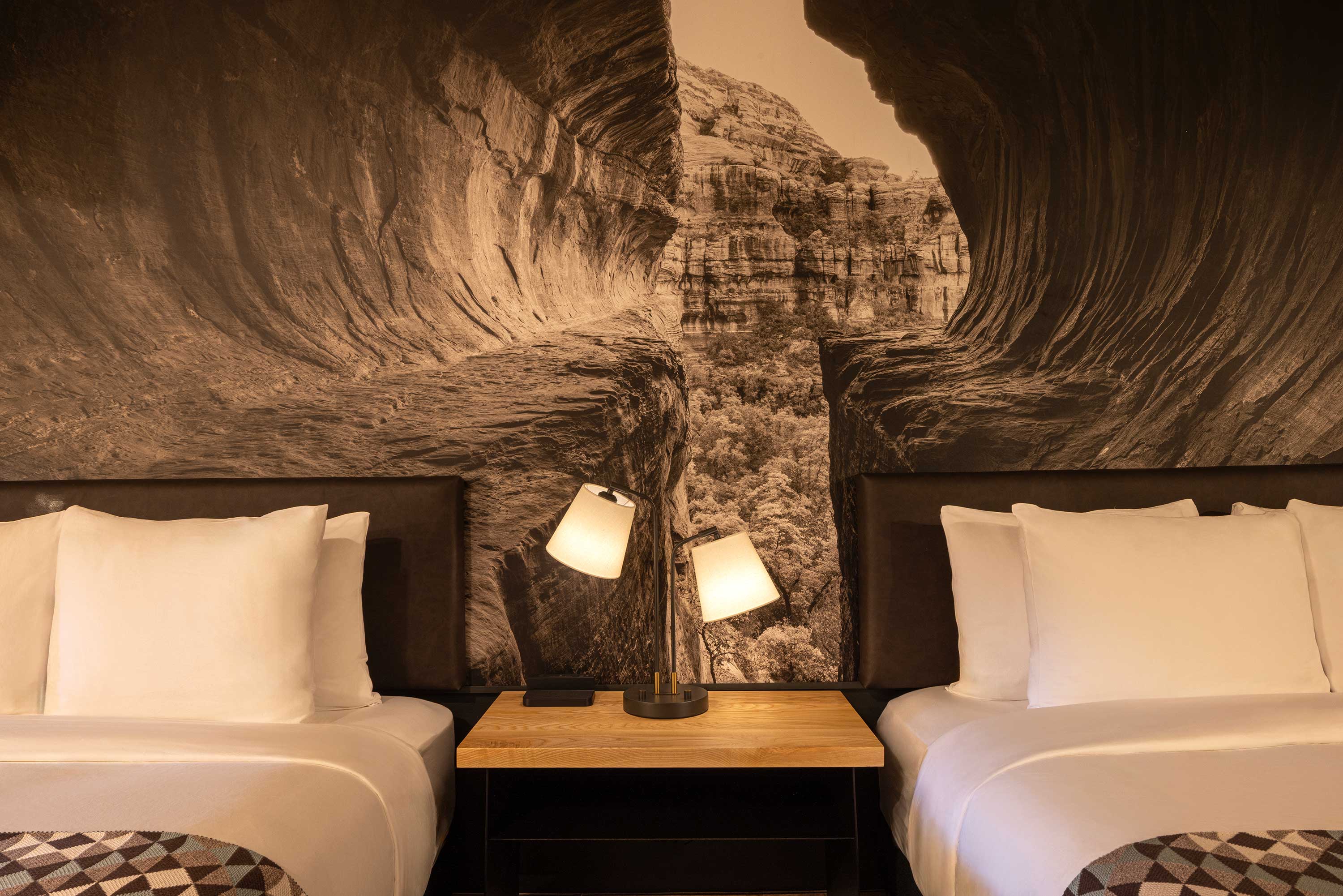 Two neatly made beds with white pillows and linens separated by a wooden nightstand with a double lamp, set against a wall mural of a rocky canyon landscape. Mountain Modern Motel Sedona