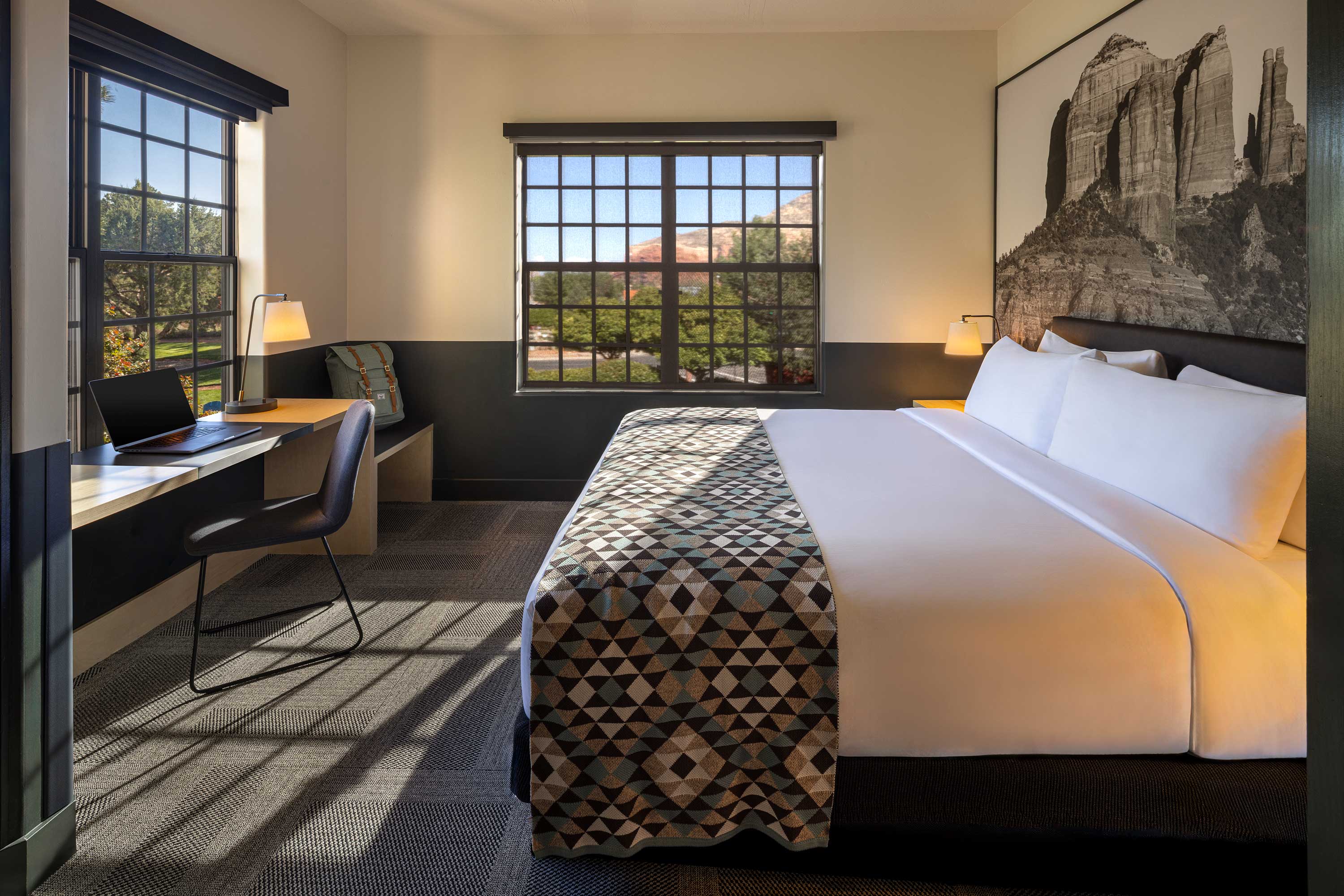 Modern hotel room with a large bed, geometric patterned runner, desk with laptop, and windows showing greenery outside. Mountain Modern Motel Sedona