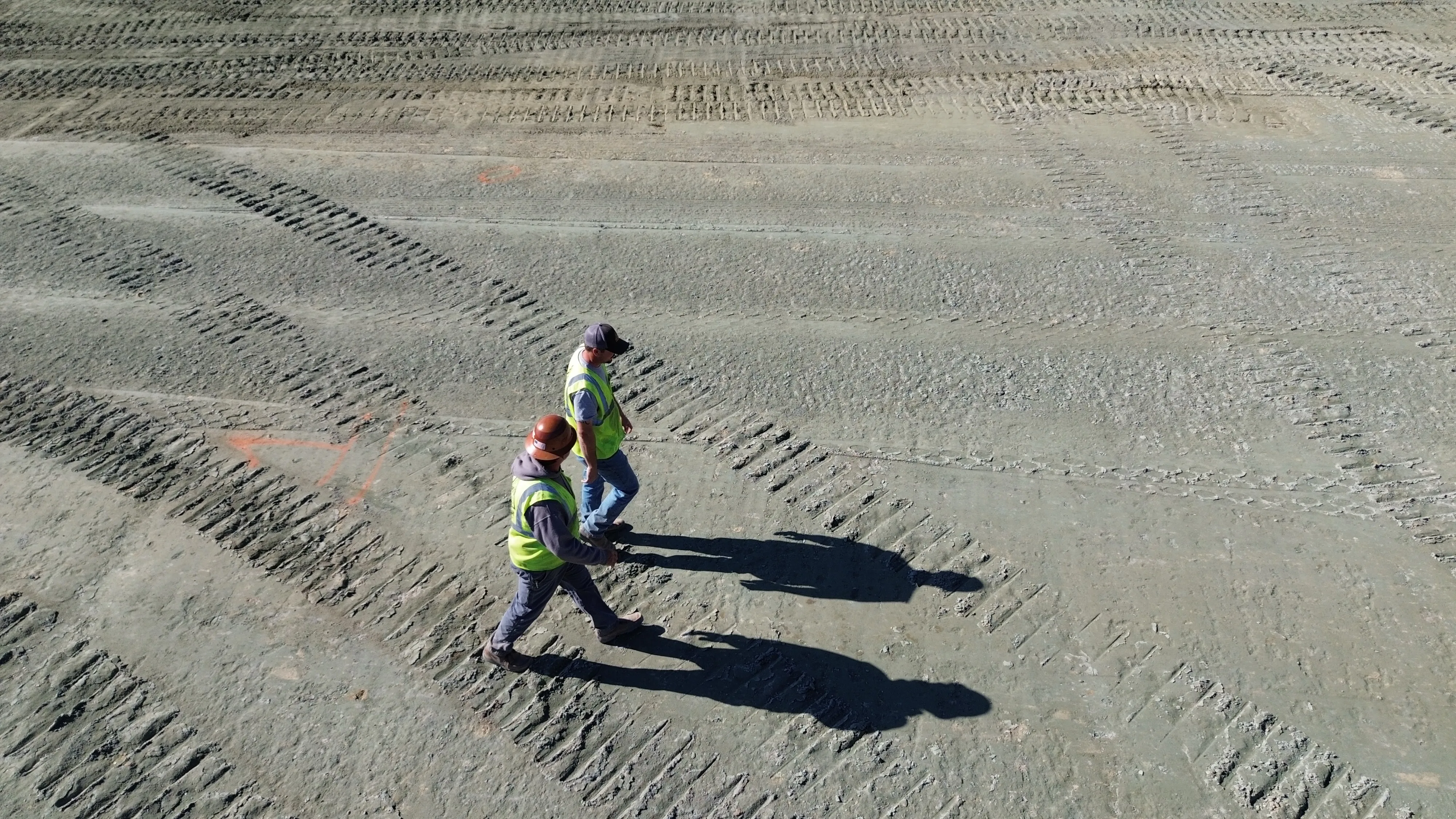 Aerial view of two construction workers in high-visibility vests inspecting furrowed dirt site.