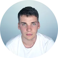 Portrait of a young man with short dark hair and blue eyes wearing a white t-shirt against a light gray background.