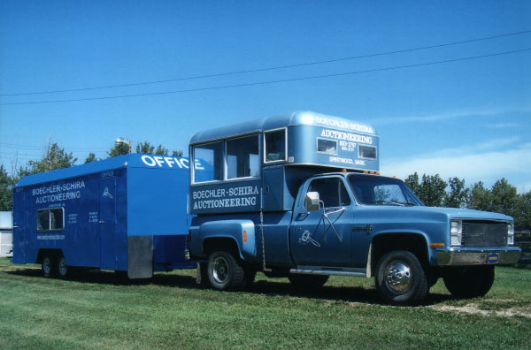 Blue auctioneering truck with attached trailer parked on green grass under a clear blue sky.