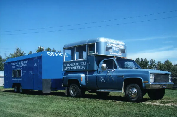 Blue auctioneering truck with attached trailer parked on green grass under a clear blue sky.