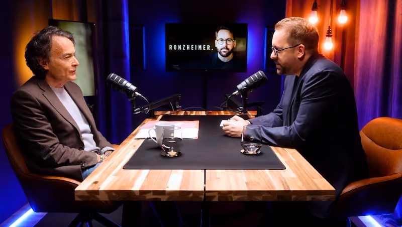 Zwei Männer sitzen an einem Holztisch in einem Podcaststudio mit Mikrofonen, einer Tasse und Gläsern, während im Hintergrund ein Bildschirm mit der Aufschrift „RONZHEIMER.“ zu sehen ist.