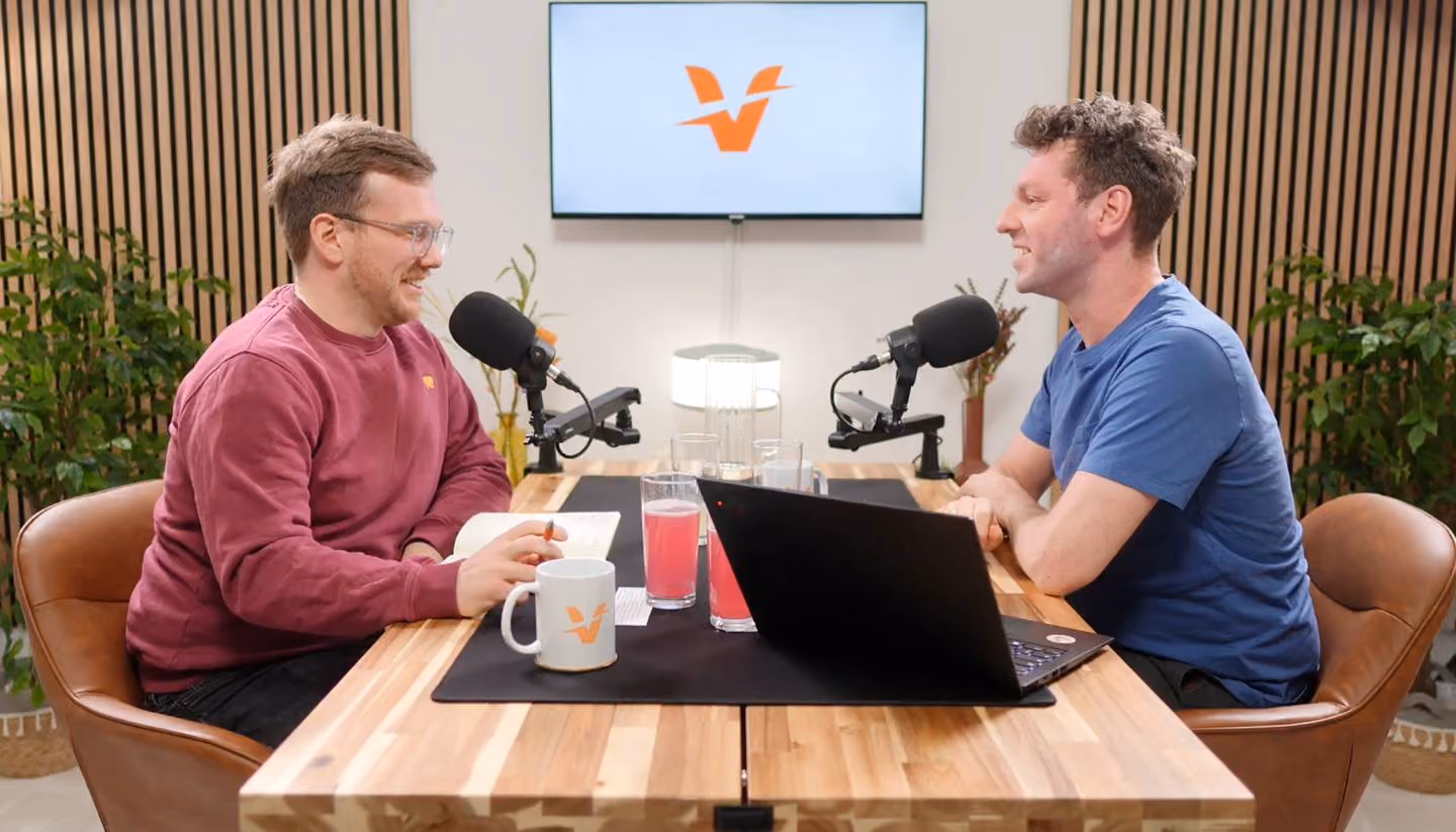 Zwei Männer sitzen an einem Holztisch mit Mikrofonen und nehmen einen Podcast auf, mit Laptop und Getränken vor sich sowie einem Bildschirm im Hintergrund, der ein orangefarbenes Logo anzeigt.