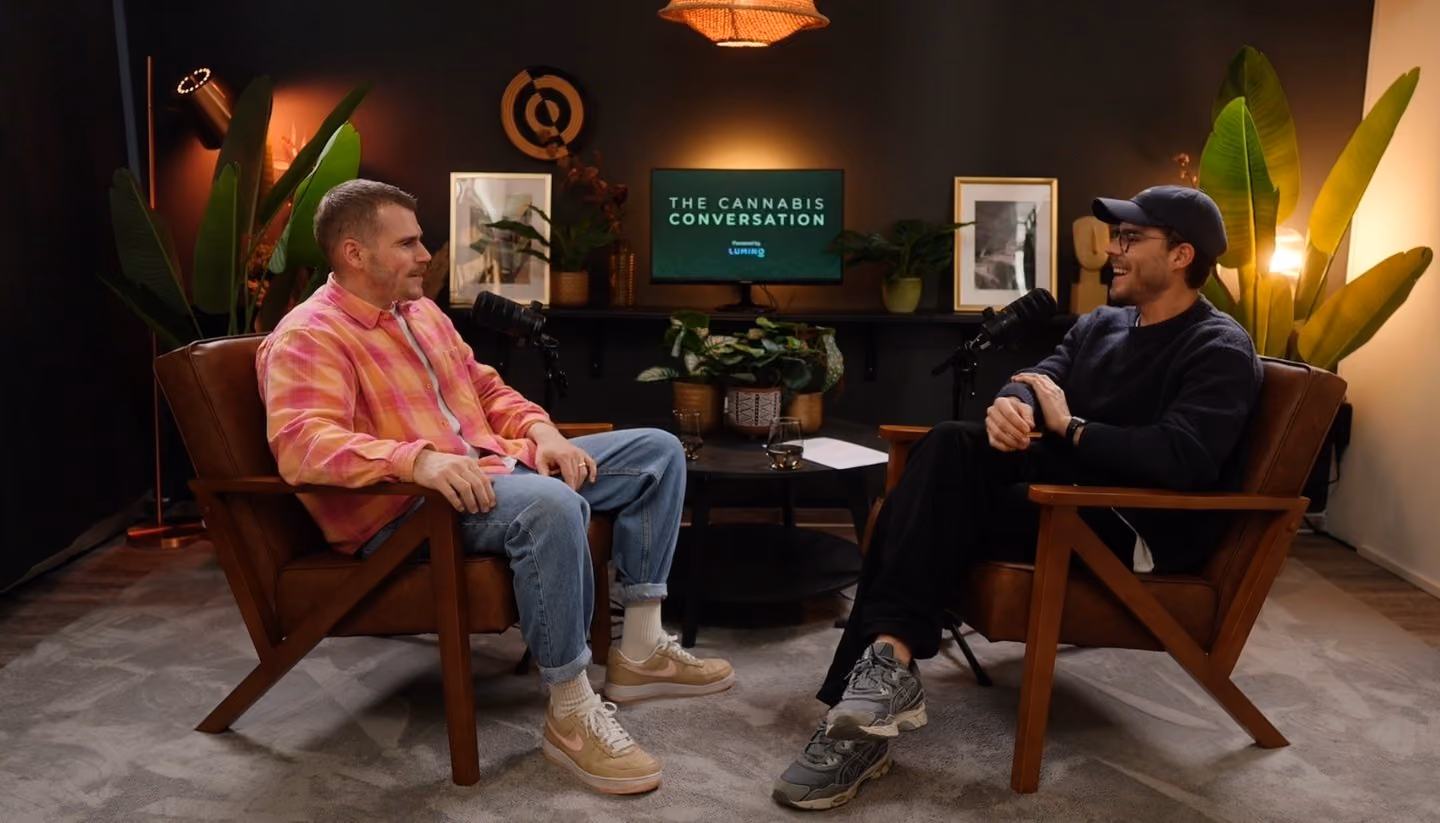 Zwei Männer sitzen in braunen Sesseln und führen ein Gespräch in einem Studio-Setup mit Mikrofonen, während im Hintergrund ein Bildschirm den Podcast „The Cannabis Conversation“ anzeigt.