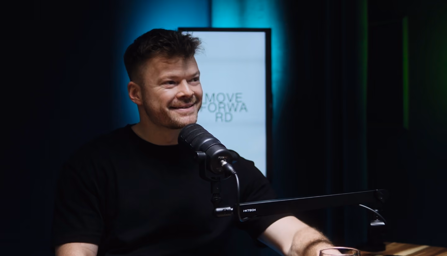 Lächelnder Mann spricht in ein Mikrofon in einem gedämpft beleuchteten Podcaststudio, während im Hintergrund ein „MOVE FORWARD“-Schild zu sehen ist.