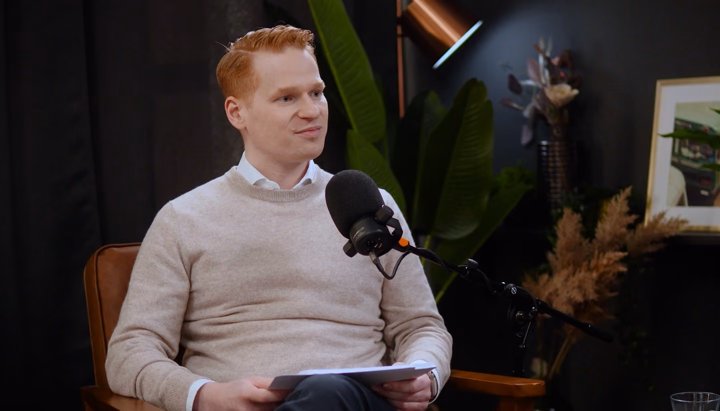 Mann in einem beigen Pullover sitzt auf einem Stuhl, hält Papiere in der Hand und spricht in ein Mikrofon in einem Studio.