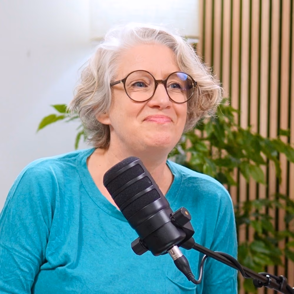 Frau mit kurzen Haaren und runder Brille spricht in ein schwarzes Mikrofon, in einem Studio mit grünen Pflanzen im Hintergrund.