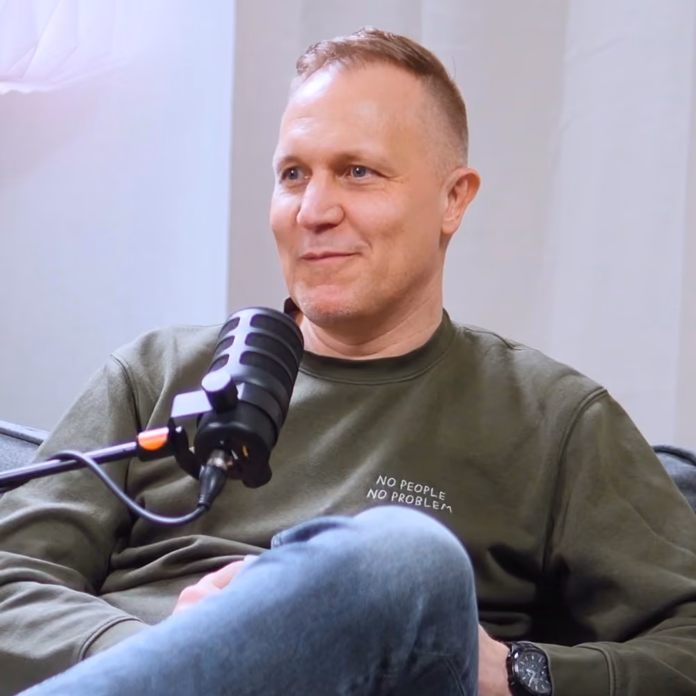 Mann in einem olivgrünen Sweatshirt mit der Aufschrift „NO PEOPLE NO PROBLEM“, der während einer Podcastaufnahme in ein Mikrofon spricht.